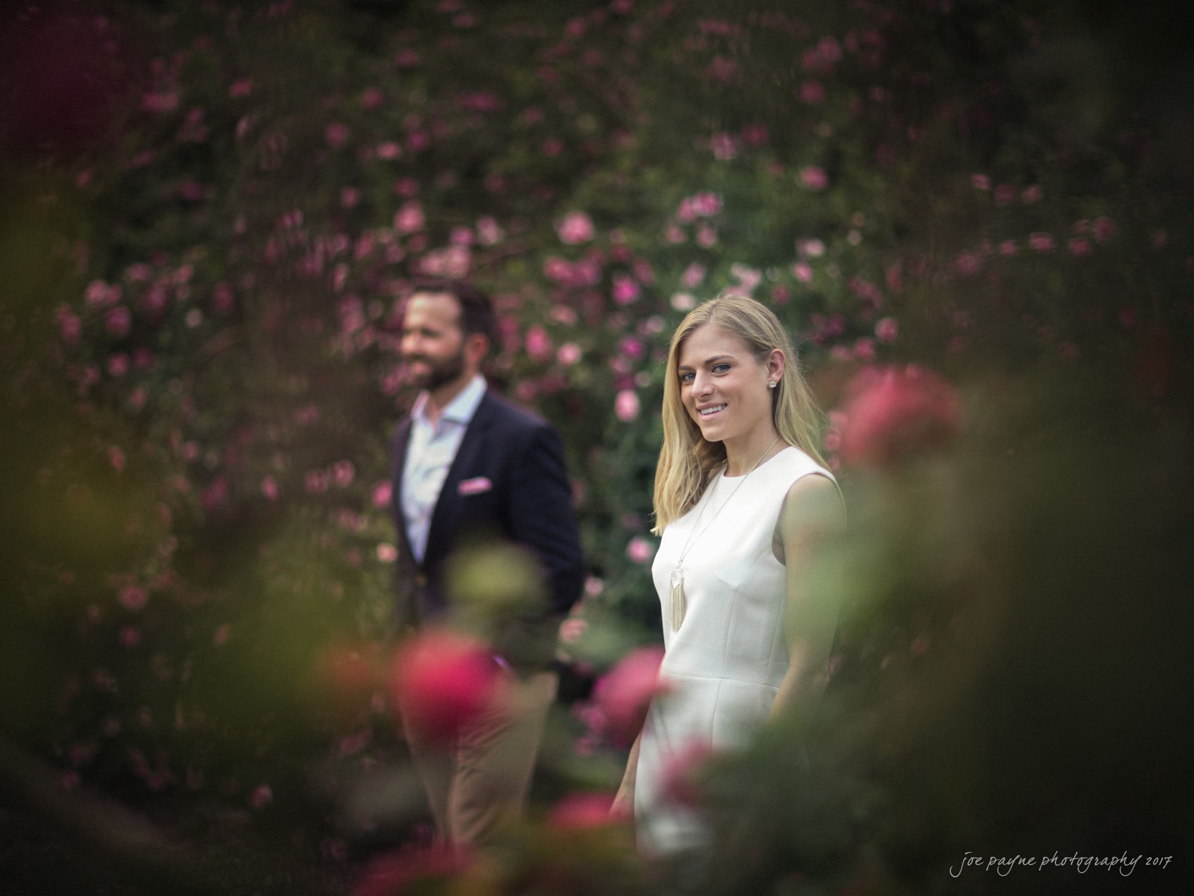 duke gardens engagement photography - margo & patrick A woman in a white dress stands smiling among pink flowers. In the blurred background, a man in a dark jacket and beige pants is also smiling. The scene is framed by vibrant, blooming flowers.