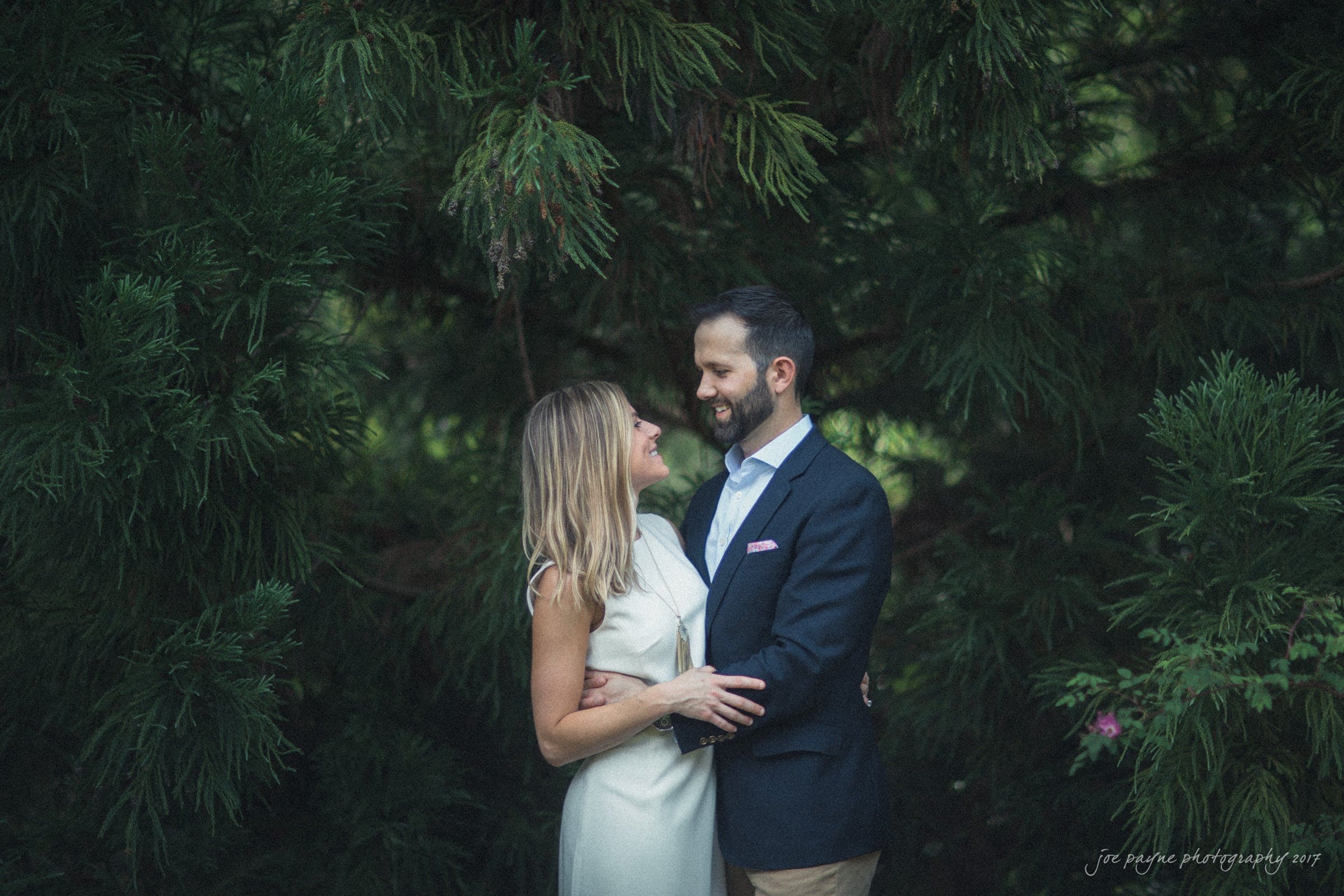 duke gardens engagement photography - margo & patrick A couple stands embraced in front of lush green foliage. The woman wears a white dress, and the man is in a navy blazer. They gaze at each other, smiling warmly.