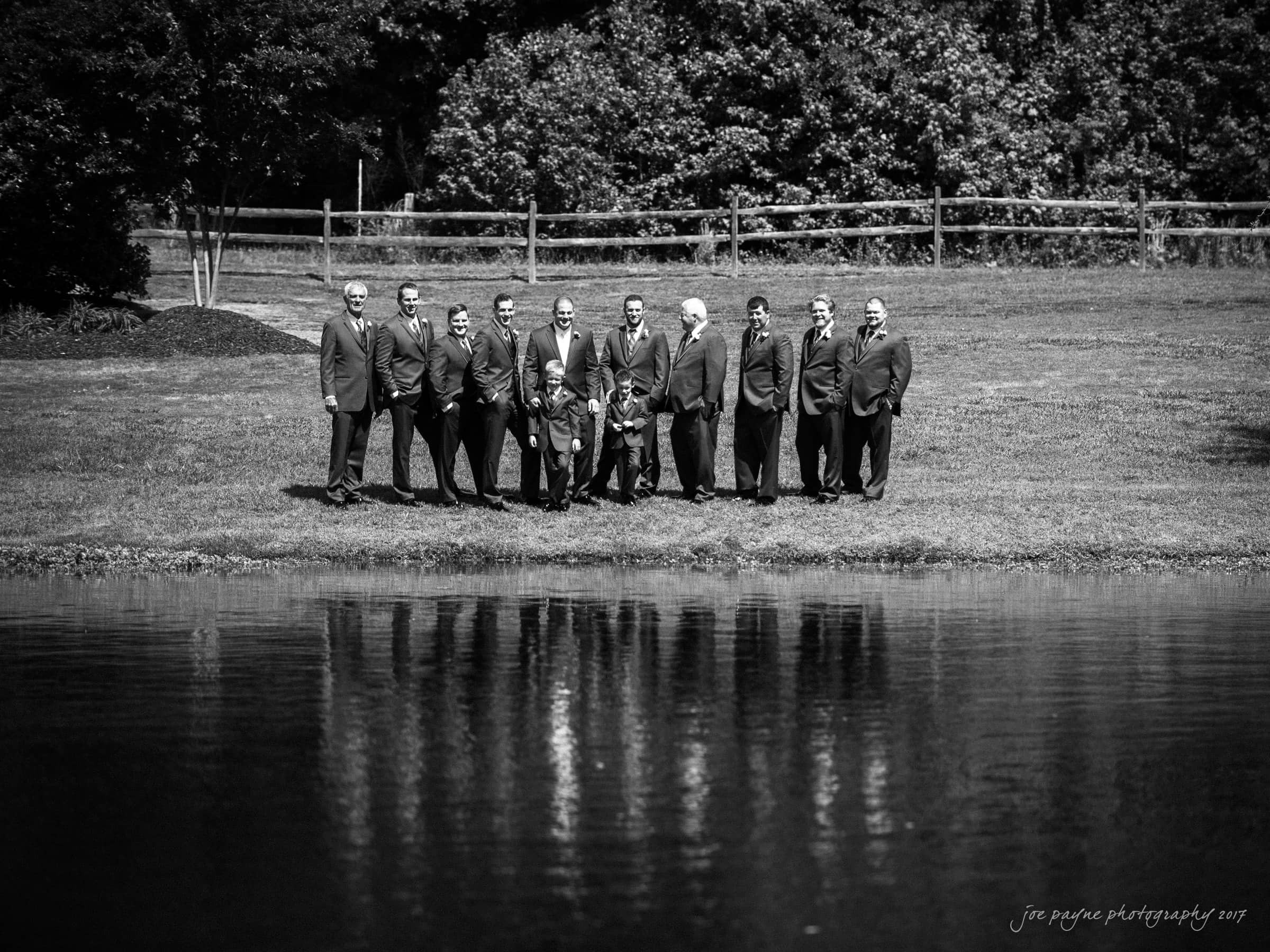 A group of people in formal attire stand together on a grassy area beside a pond. Trees and a wooden fence are in the background. The reflection of the group is visible in the water.
