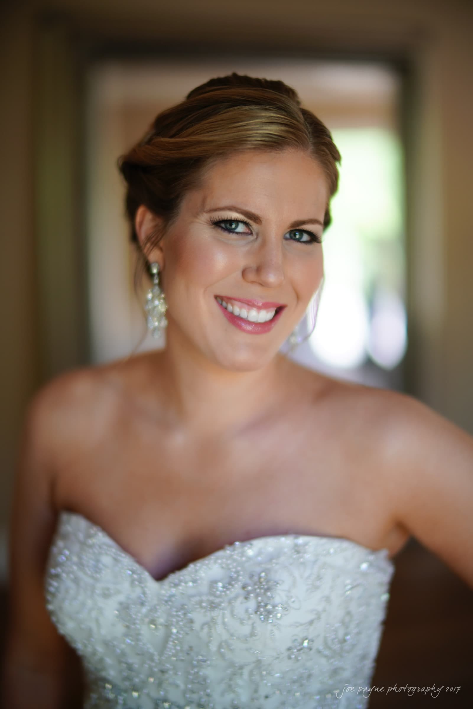 A smiling bride in a strapless white wedding dress adorned with intricate beadwork, standing indoors. She has her hair elegantly styled up, and she wears sparkling earrings. The background is softly blurred.