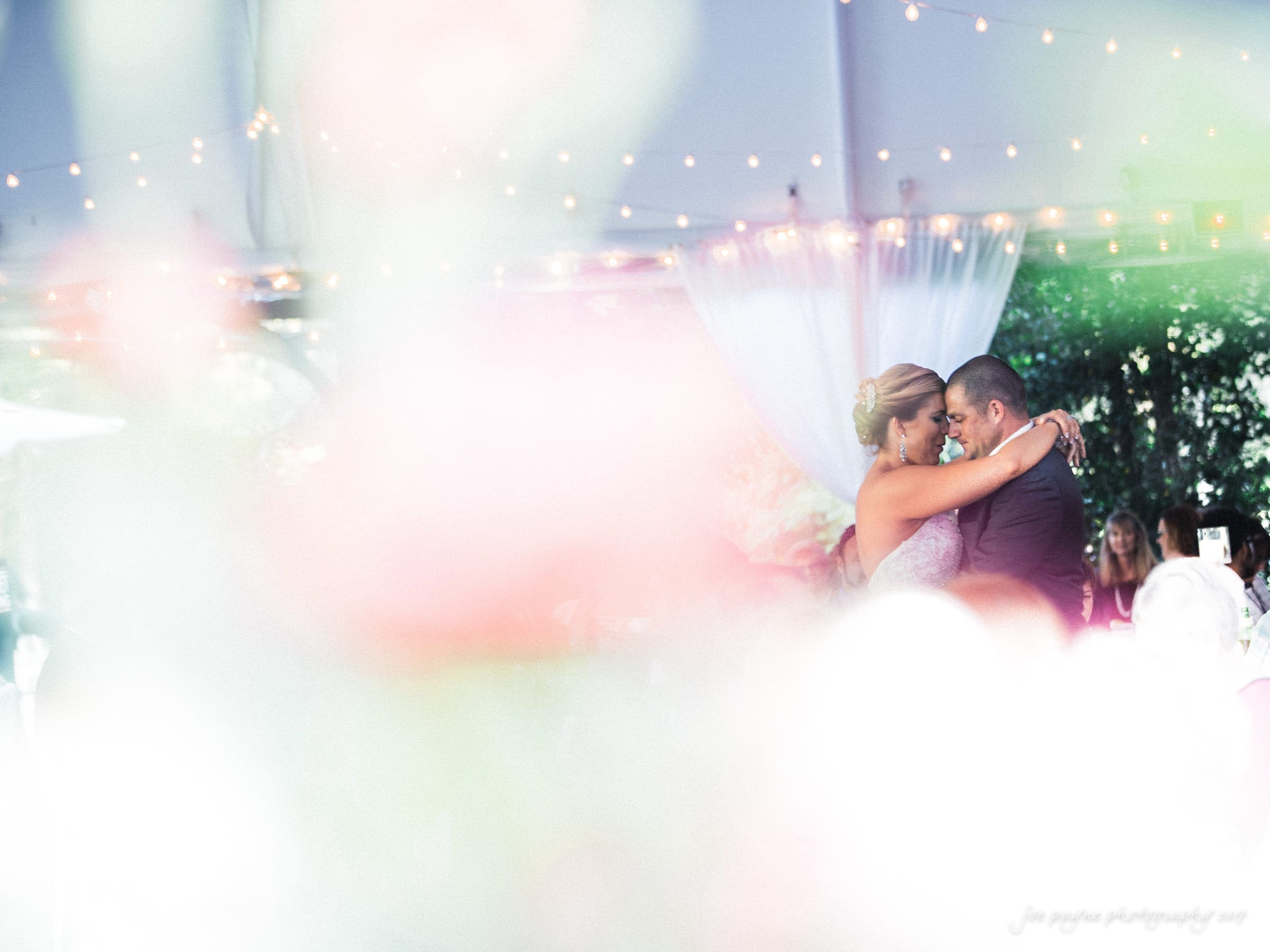 A couple shares a romantic dance under soft string lights. The background is softly blurred, highlighting their embrace. The atmosphere is intimate and dreamy, framed by gentle drapery.