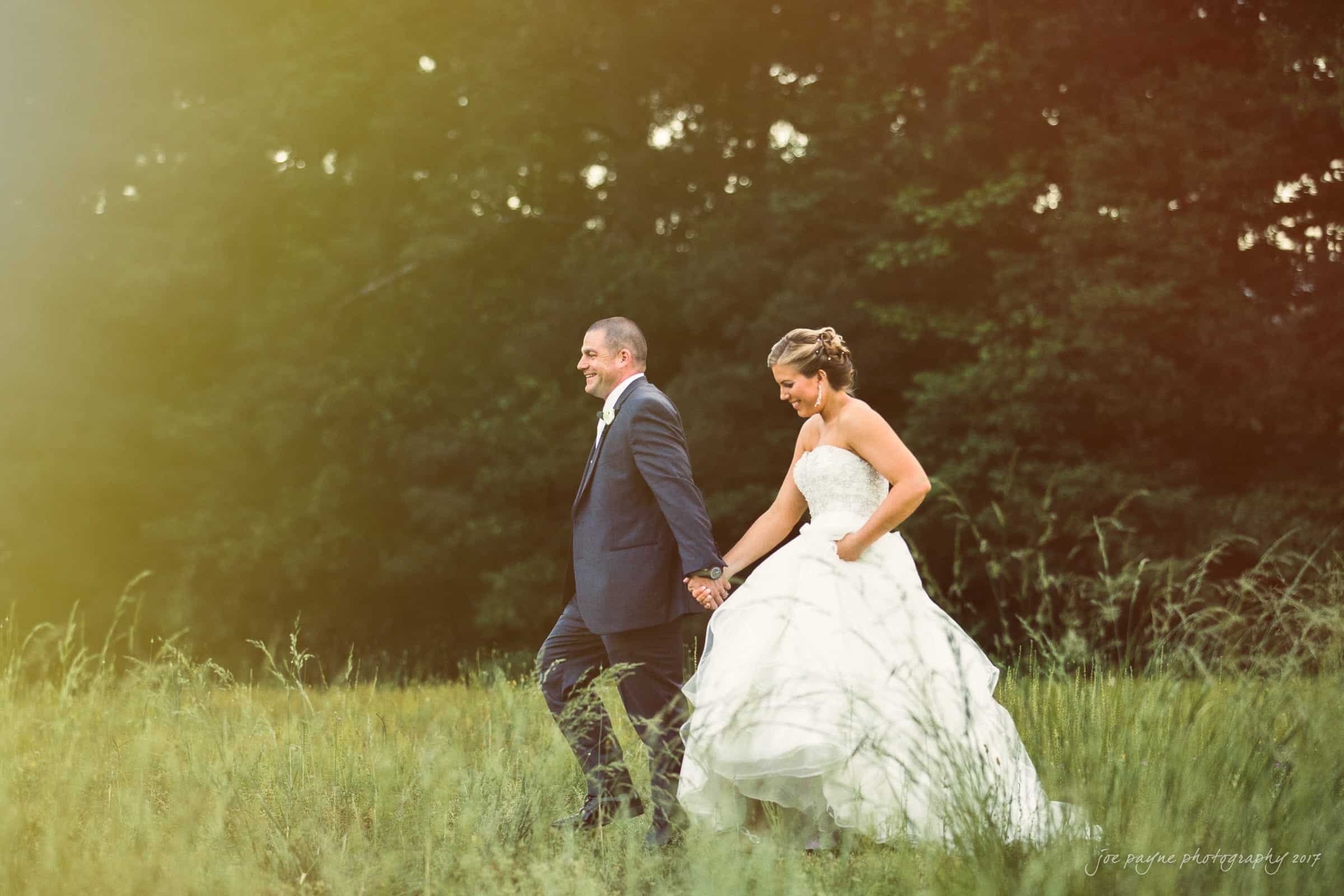 A couple in wedding attire walks hand in hand through a grassy field, with sunlight filtering through trees in the background. The groom is wearing a dark suit, and the bride is in a white gown.