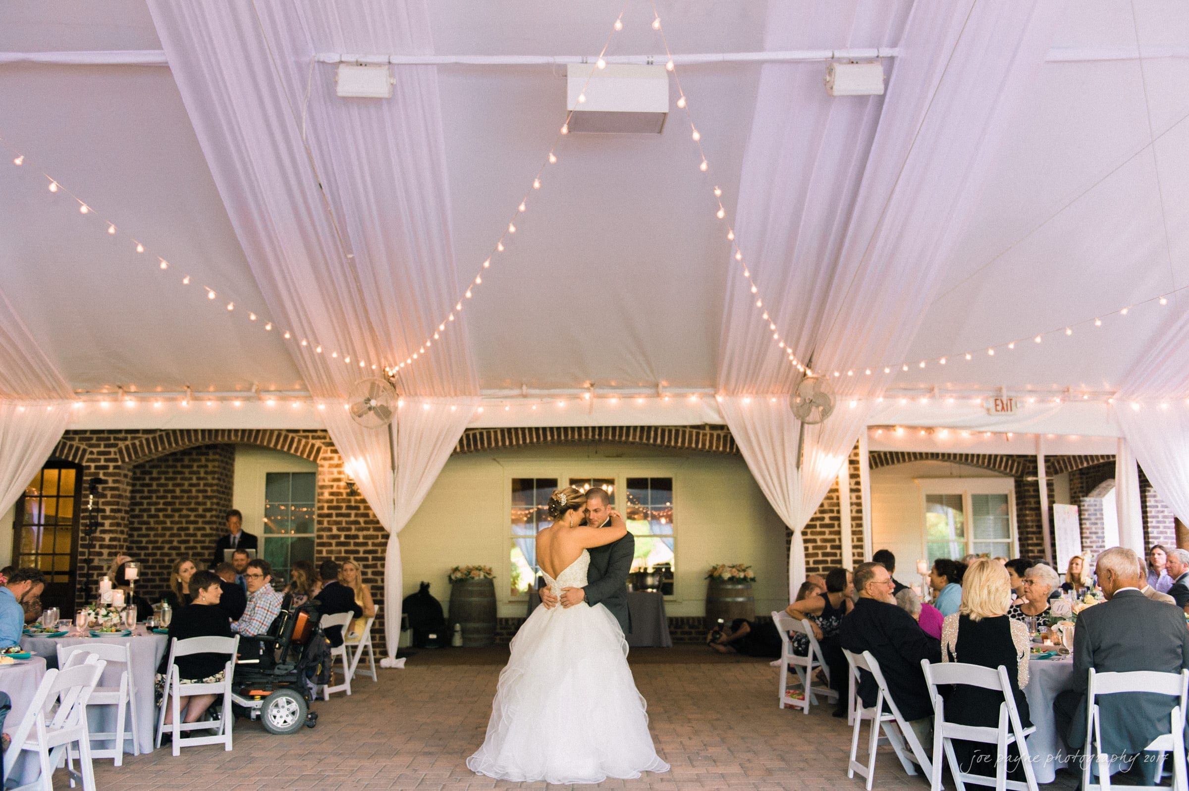 A couple enjoys their first dance at their wedding reception under a white tent with string lights. Guests are seated at round tables, watching and celebrating in a cozy, festive atmosphere.