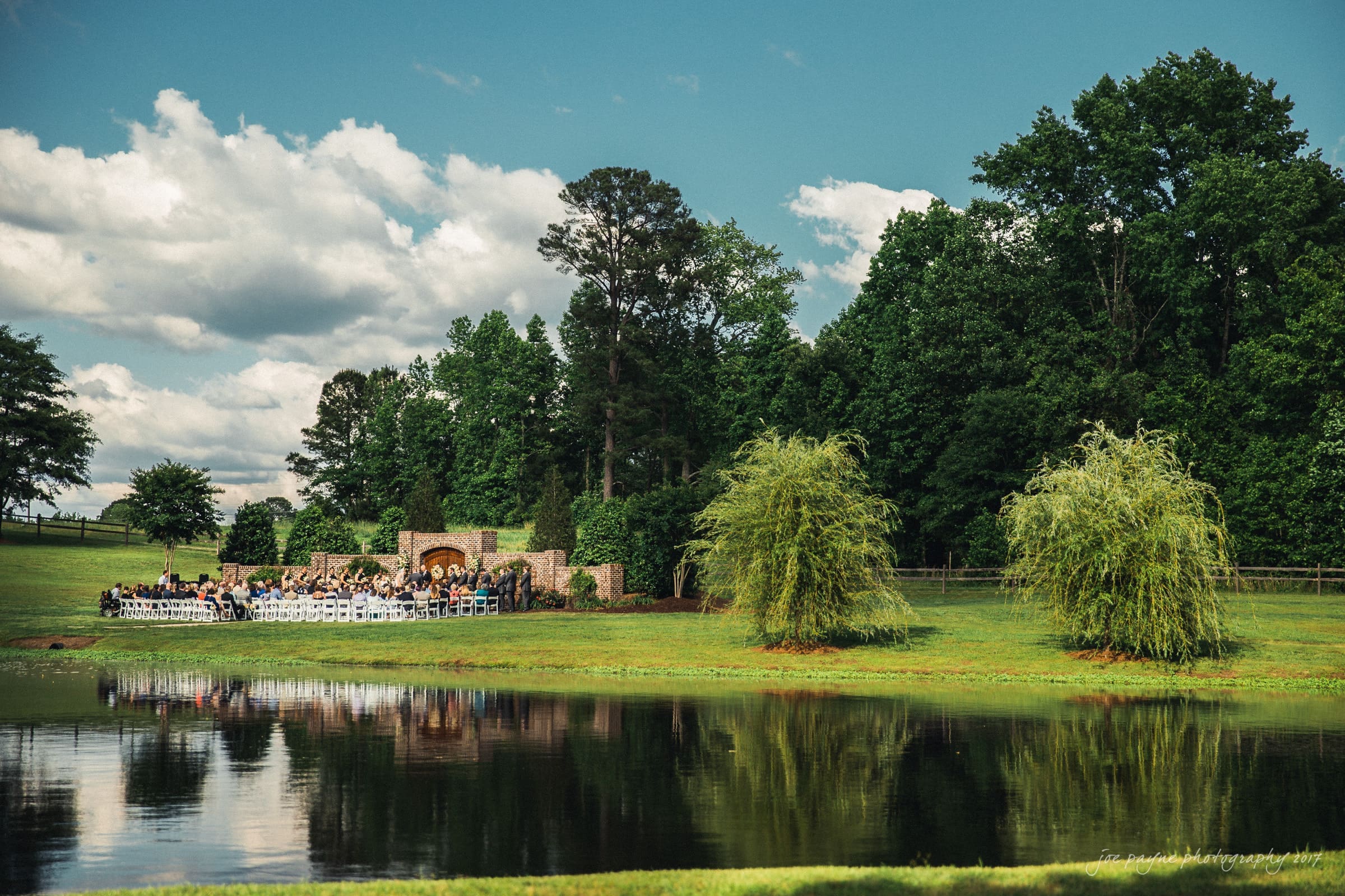 A picturesque outdoor wedding ceremony by a serene lake, surrounded by lush greenery and tall trees. Guests are seated on white chairs, facing a rustic stone backdrop under a clear blue sky with scattered clouds.