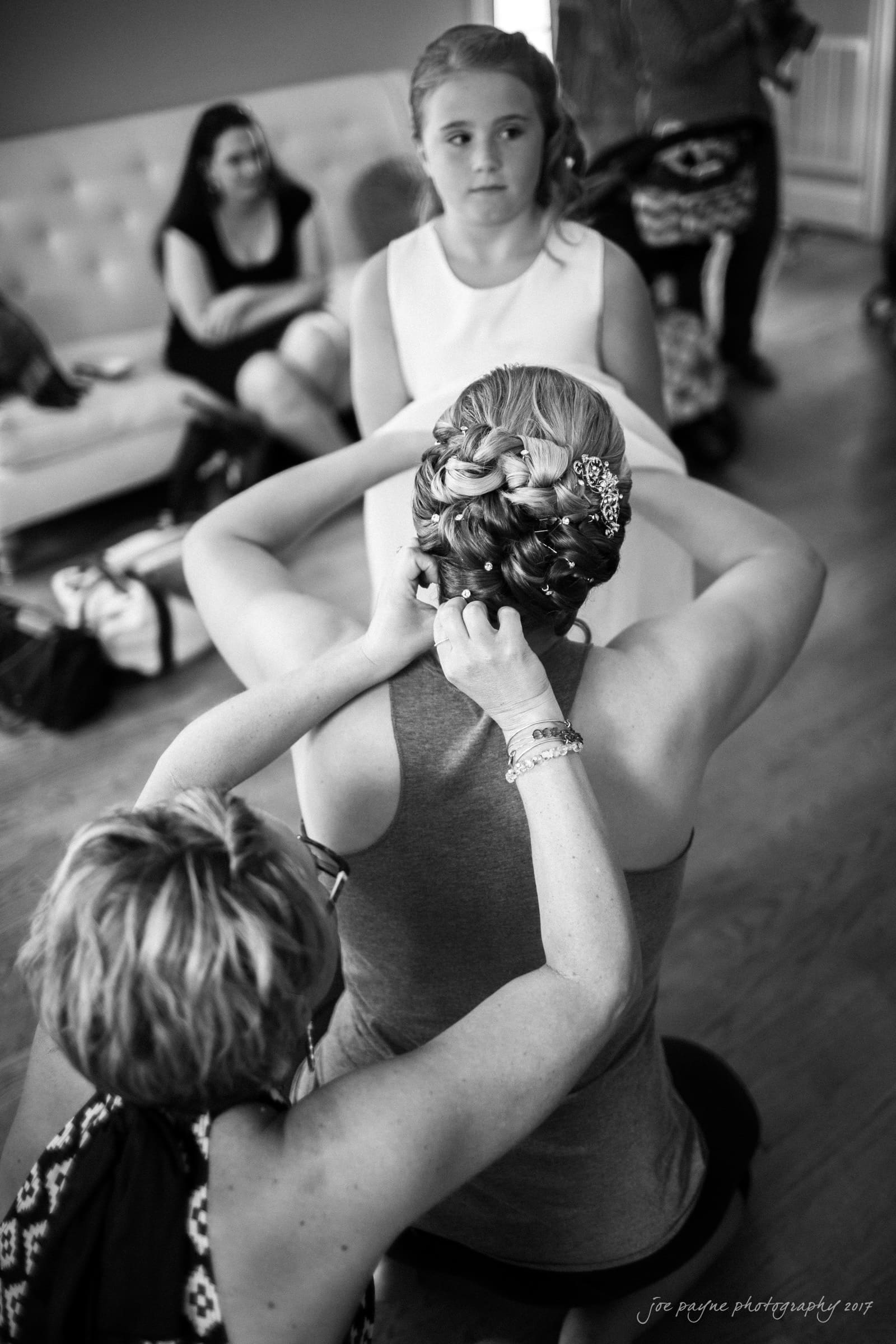A woman styles the intricate braided hair of another woman sitting in front of her, while a young girl in a white dress watches. Other people are seated on a couch in the background. The setting appears to be a preparation room.