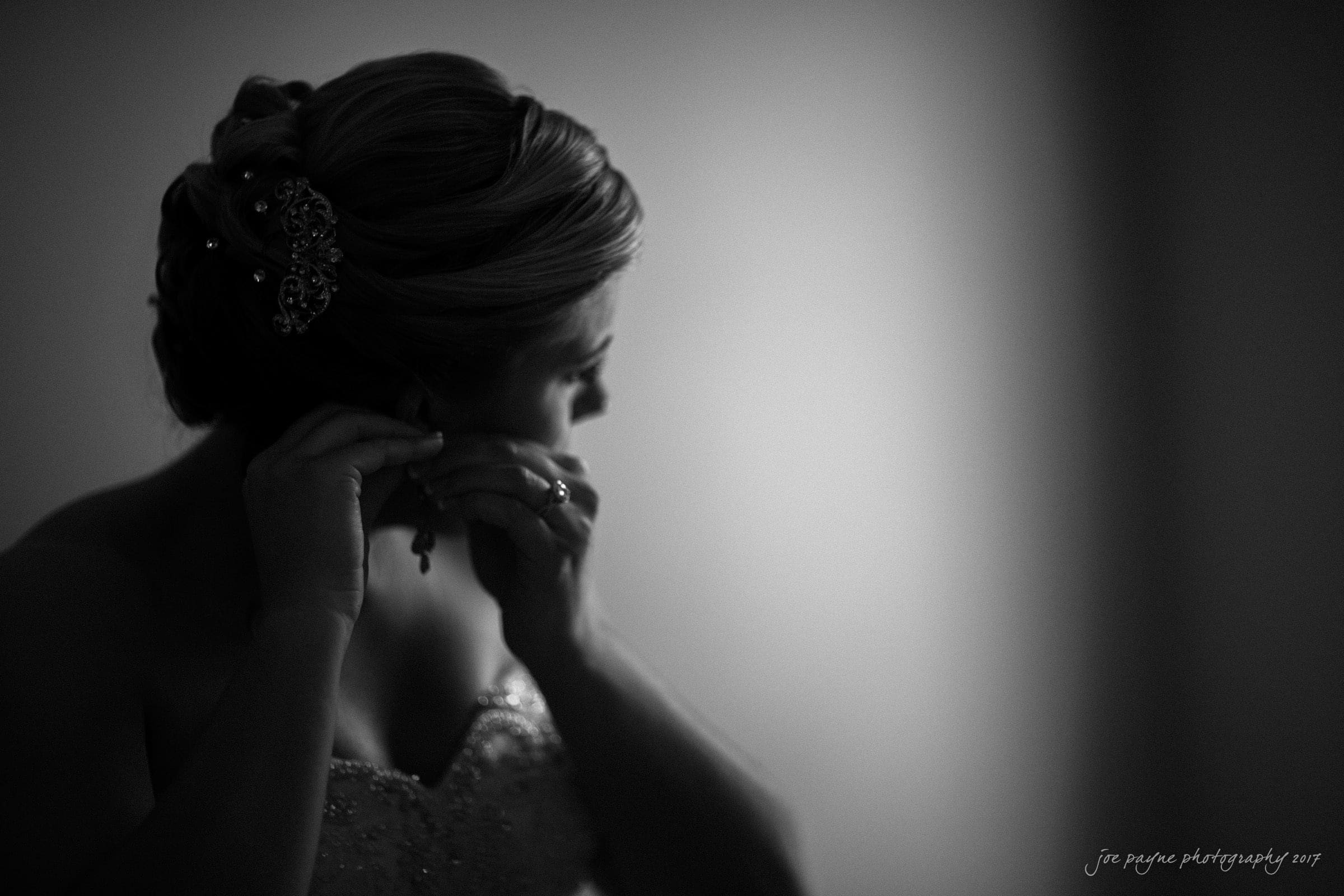 A bride, seen in silhouette, adjusts her earring. She wears a strapless gown and her hair is elegantly styled with decorative clips. The lighting is soft and focused, creating a dramatic and intimate atmosphere.