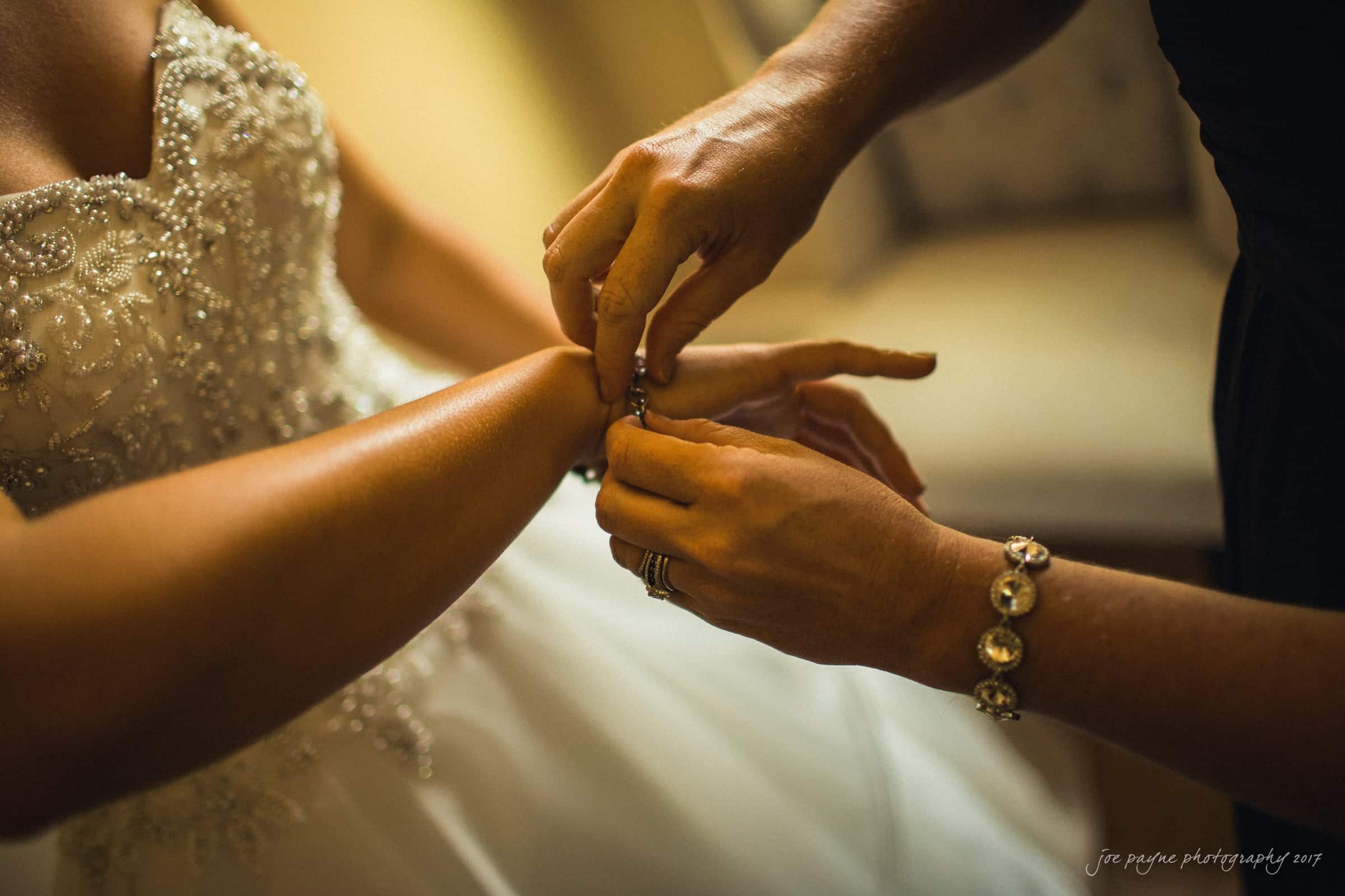 A close-up of a bride in a white, beaded wedding dress having a bracelet fastened on her wrist by another woman. The scene is warmly lit, showcasing intricate details of the dress and jewelry.