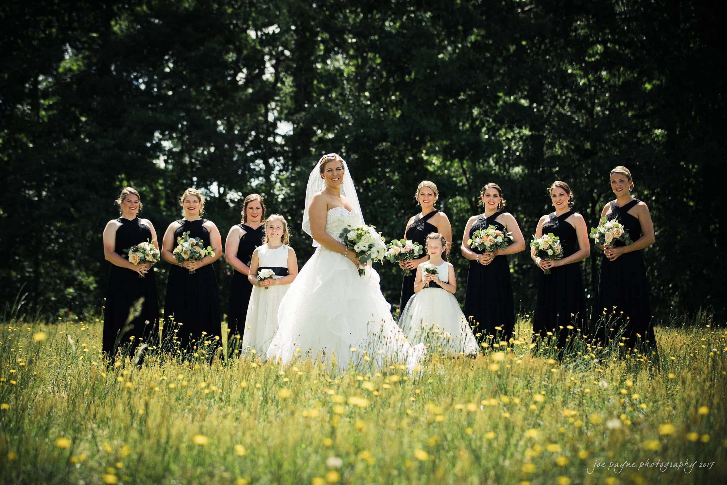 A bride in a white gown stands in a meadow surrounded by eight bridesmaids dressed in black. Each holds a bouquet of flowers. The group smiles under a clear sky with a backdrop of trees.