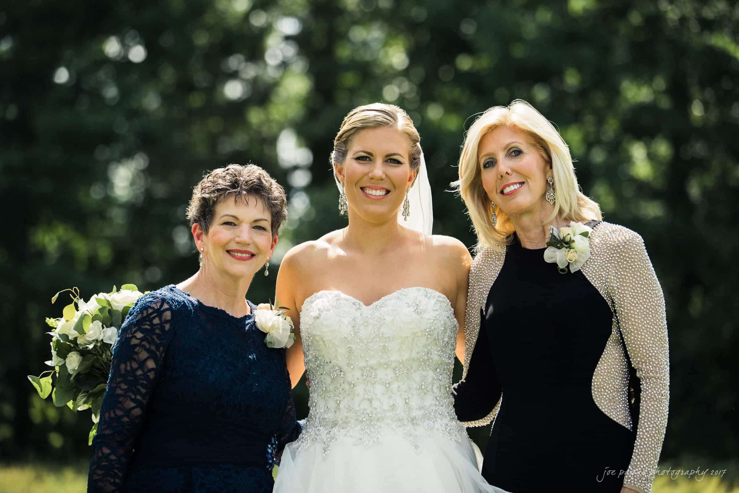 Three women smiling outdoors; the central woman in a white wedding dress, flanked by two women in elegant attire, each wearing corsages. They stand against a backdrop of lush greenery.