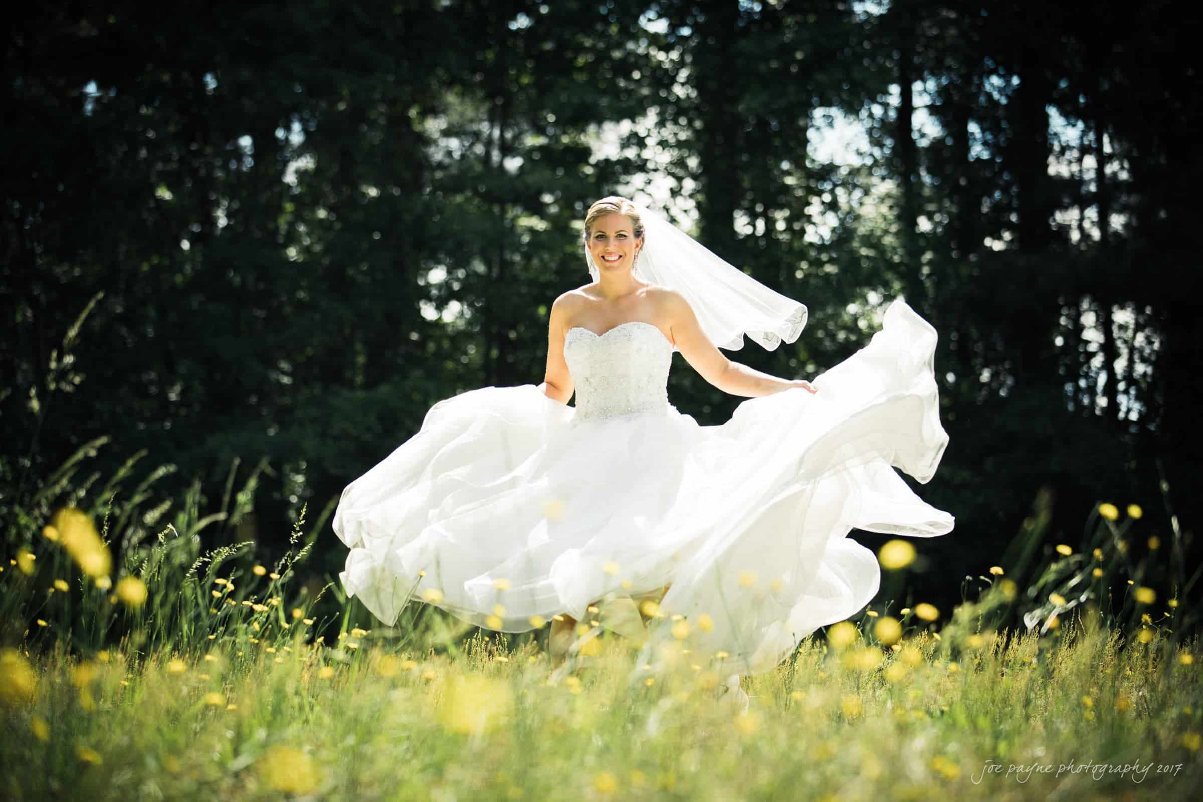 A bride joyfully twirls in a field of yellow flowers, her white gown billowing around her. Sunlight filters through the trees in the background, creating a vibrant and whimsical scene.