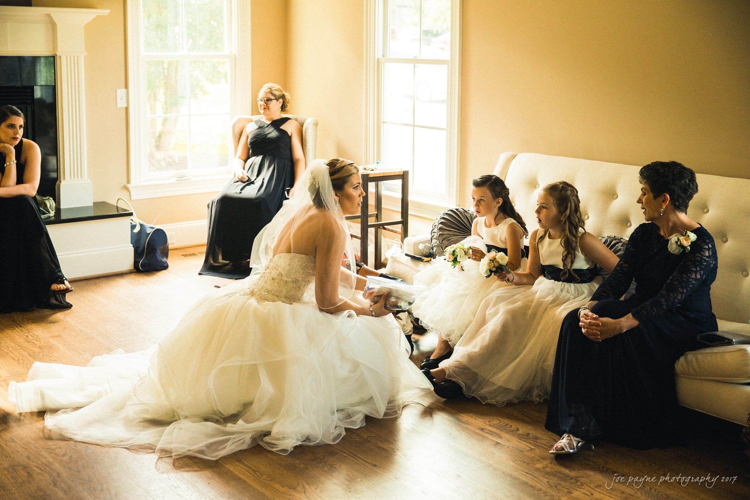 A bride in a white gown sits on a wooden floor, talking to two flower girls in white dresses who are seated on a sofa. Four women, dressed in black, are scattered around the room, some seated and one standing by a window.