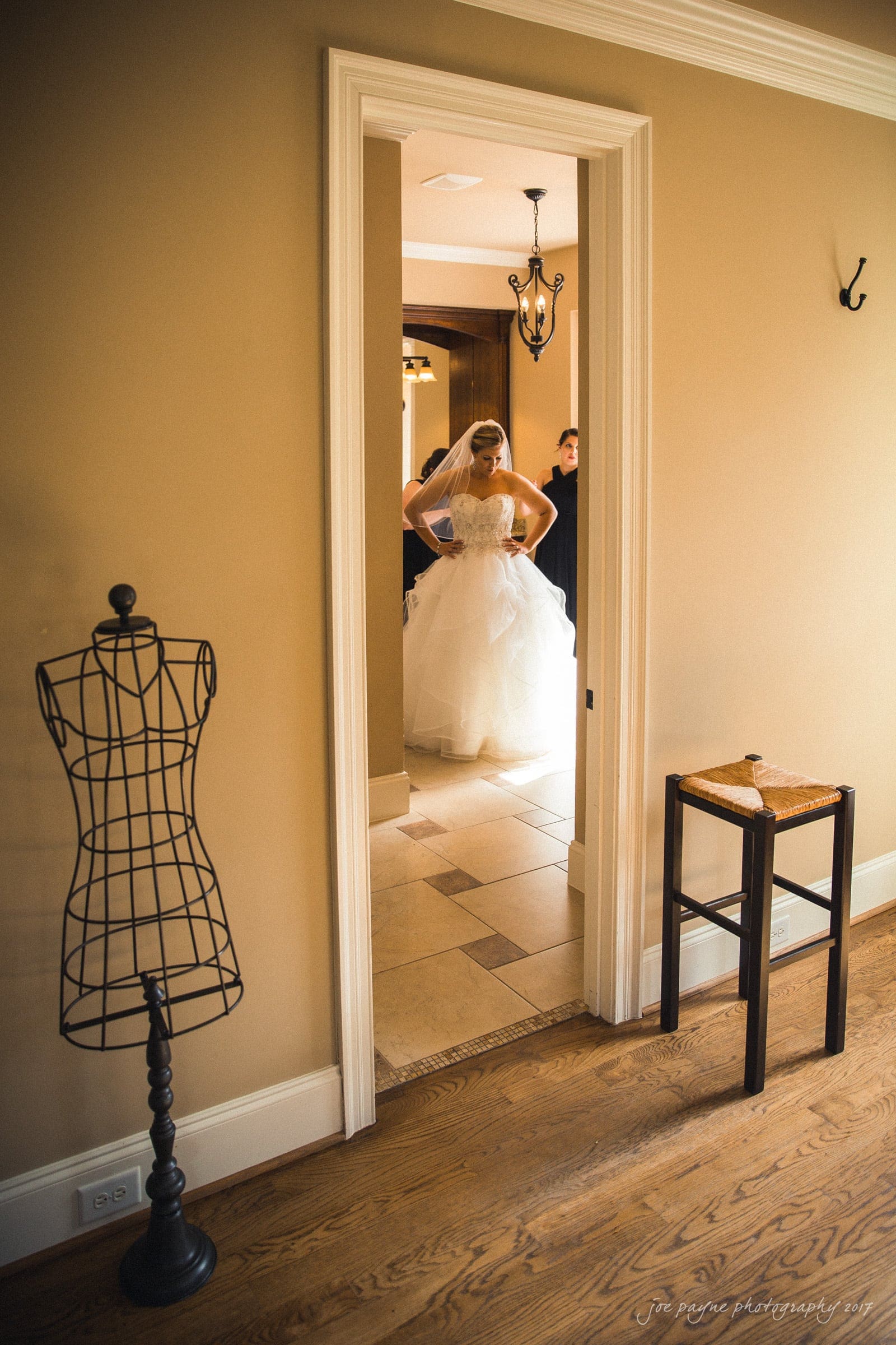 A bride in a white gown stands in a hallway, framed by a doorway. There is a decorative dress form and a small wooden table in the foreground. The setting is warmly lit with neutral-colored walls.