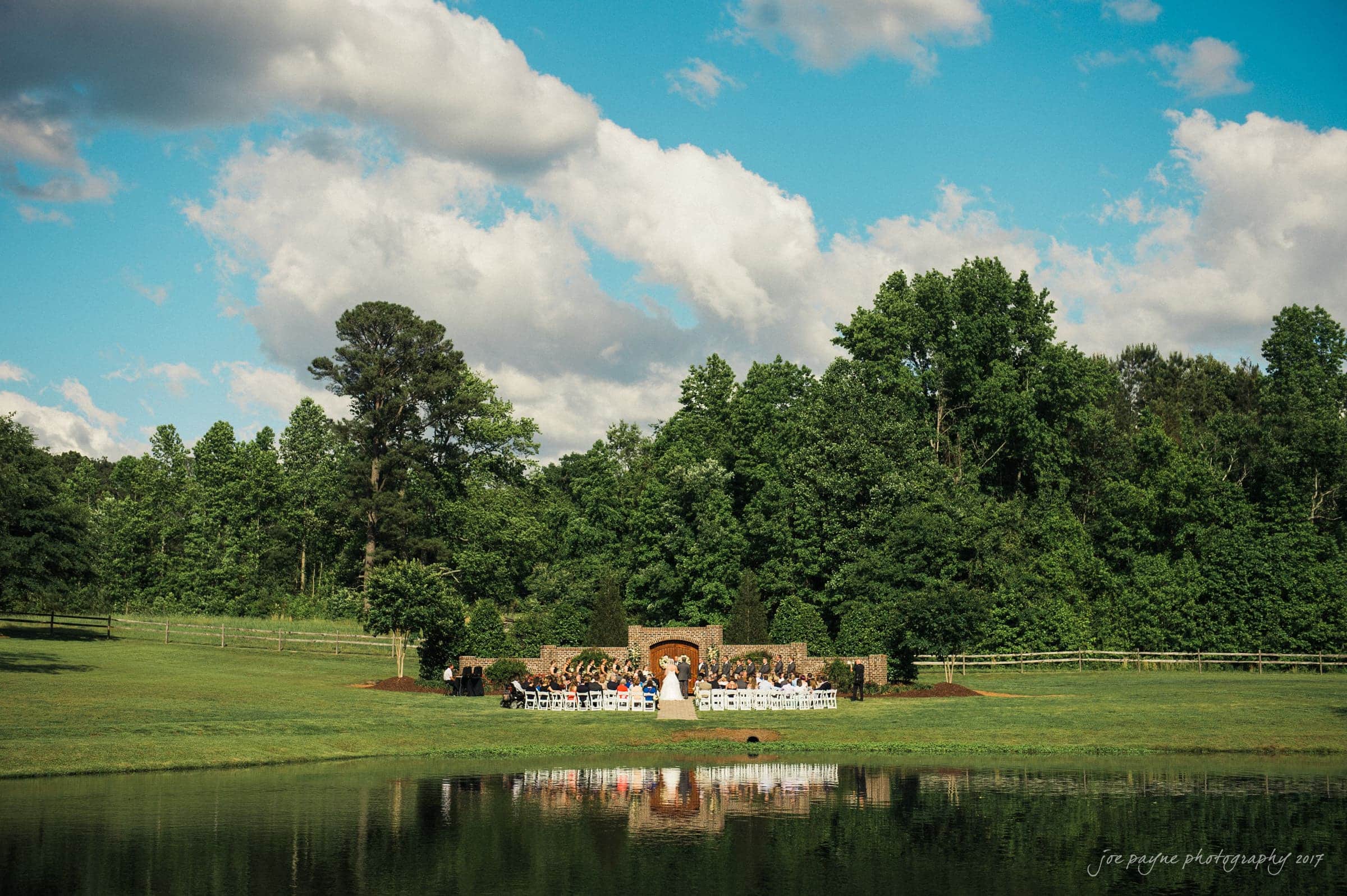 A wedding ceremony set in a lush green field with a pond in the foreground. Guests are seated on either side of an aisle leading to a floral arch, where the couple stands. Tall trees and a partly cloudy sky are in the background.