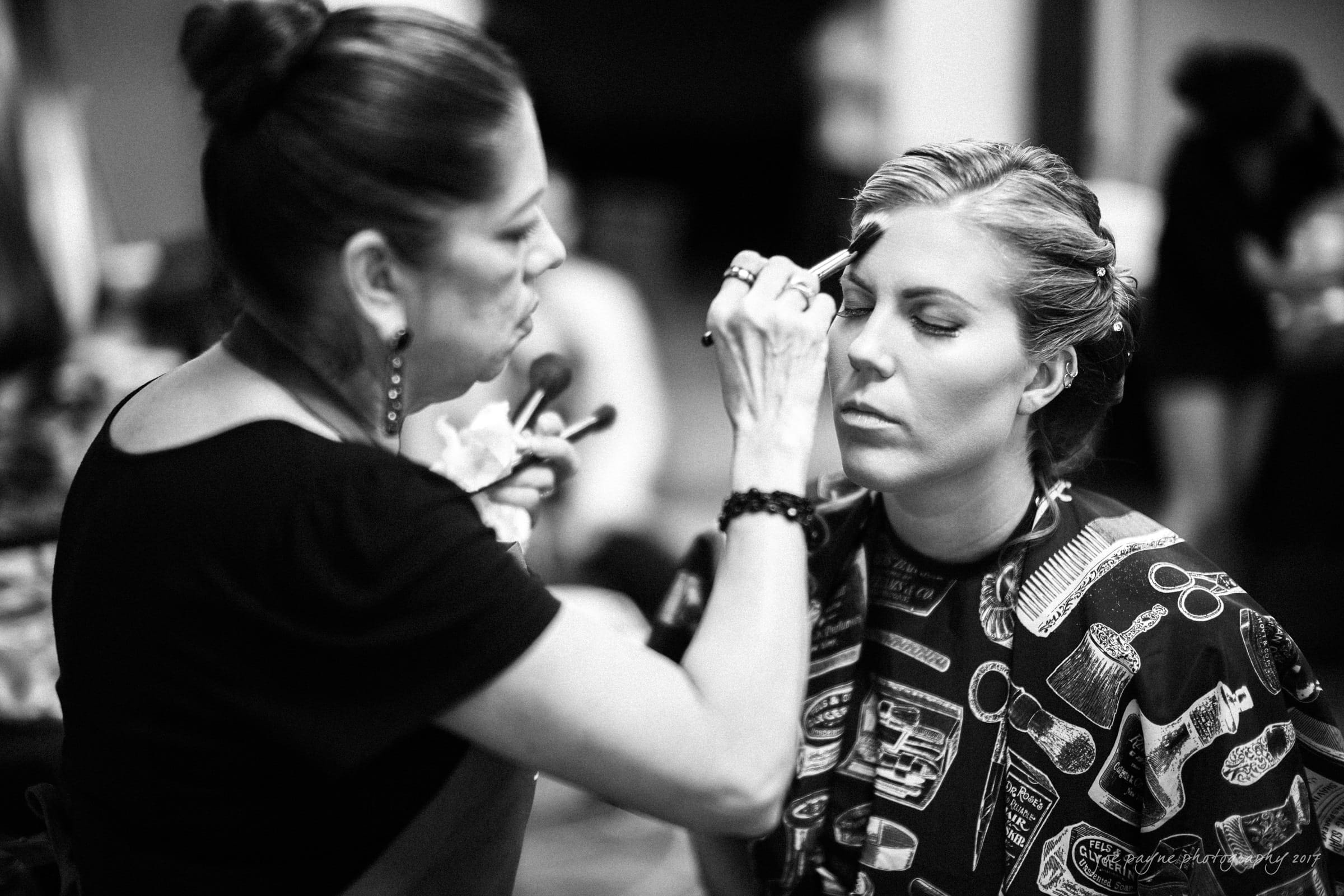 A woman with closed eyes is having makeup applied by another woman. The scene is captured in black and white, with a focus on the makeup application process. The woman receiving makeup wears a patterned cape.