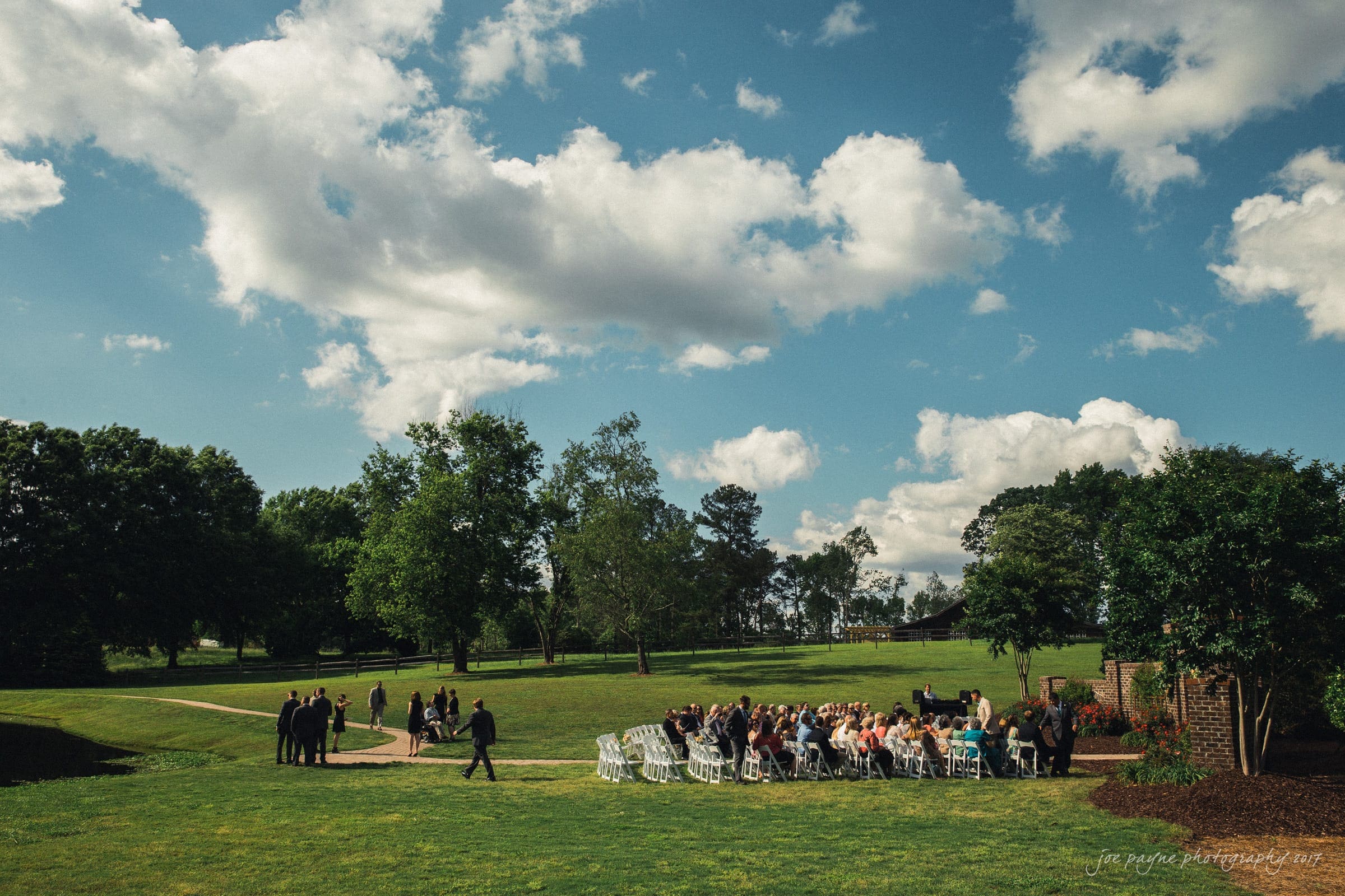 A scenic outdoor wedding ceremony with guests seated in white chairs on a grassy field. Trees and lush greenery surround the area under a blue sky with fluffy clouds. Some guests are standing, with the bride and groom in the distance.