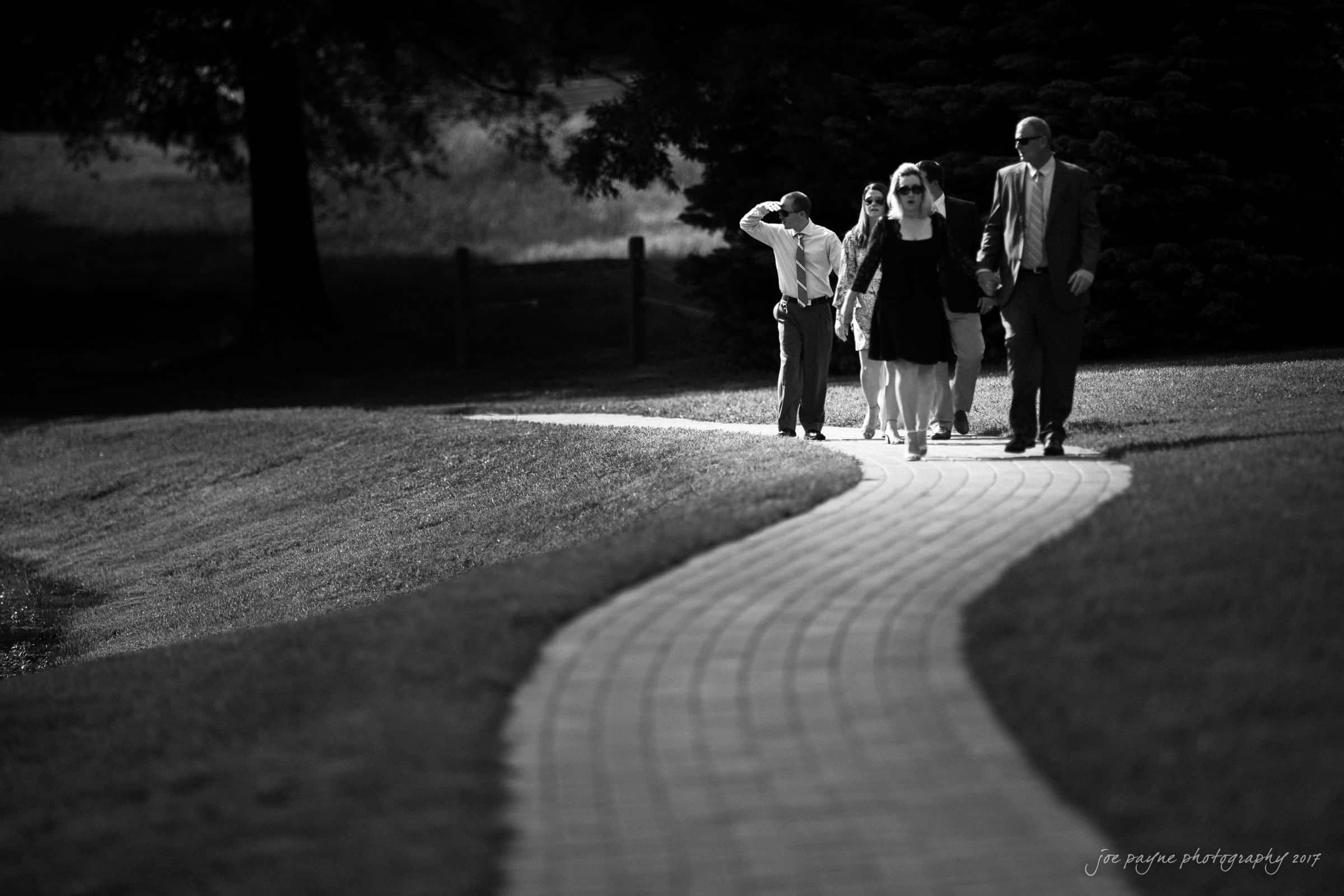 A group of people dressed formally walks along a winding brick path in a park. The scene is in black and white, with shadows cast on the grass and trees in the background.