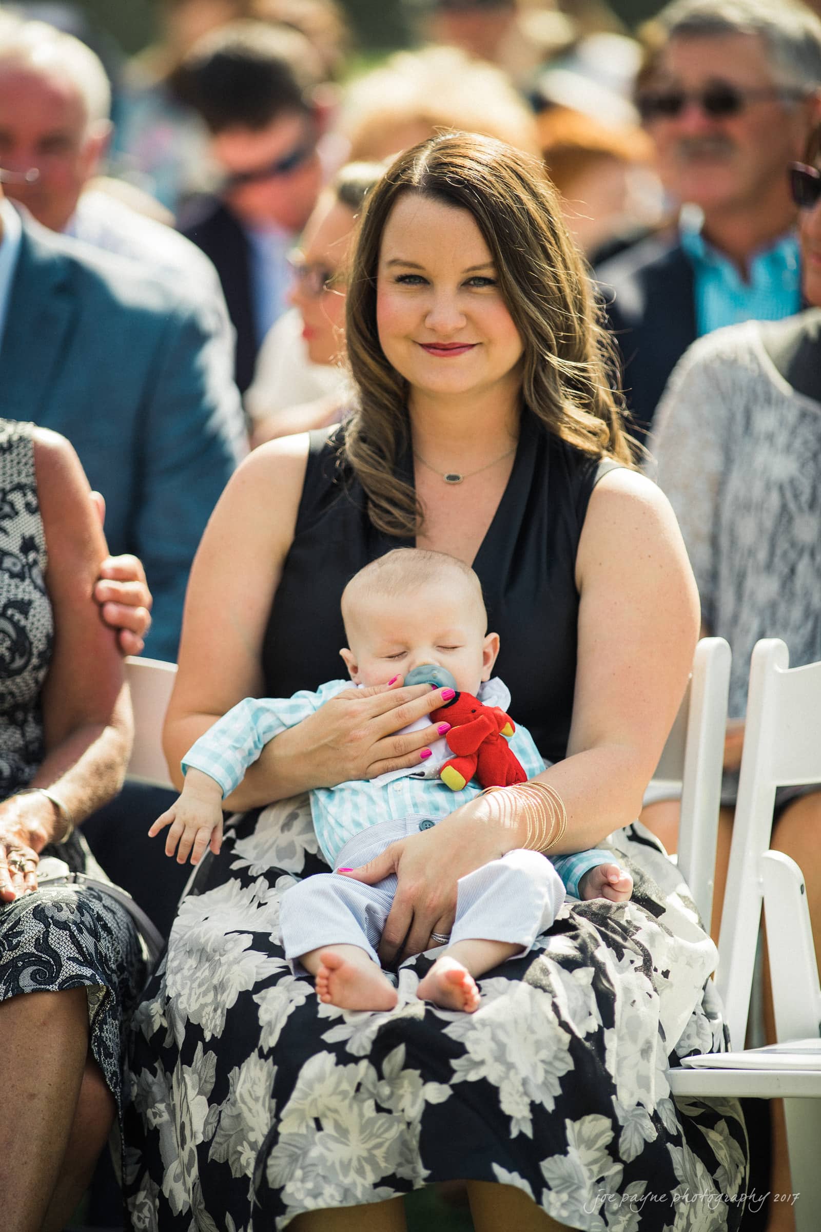 A woman sits outdoors at an event, holding a sleeping baby dressed in a blue and white outfit. She wears a black top and patterned skirt, smiling at the camera. People sit in the background.