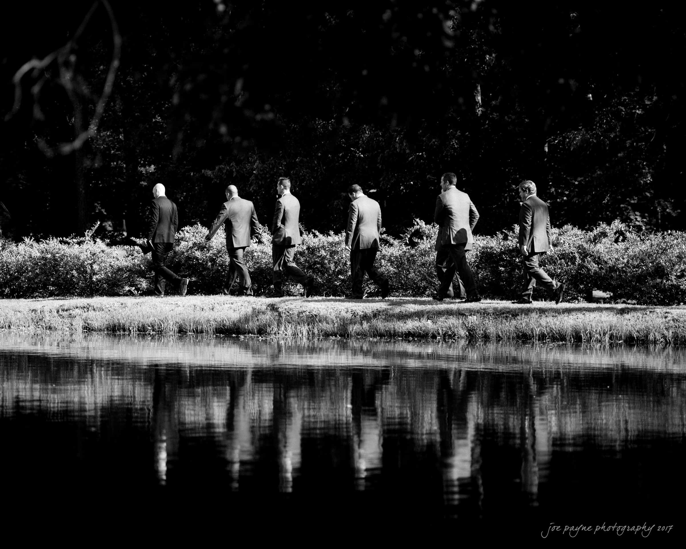 A group of seven people in suits walk along a path beside a reflective body of water. The scene is framed by trees and bushes, and the image is in black and white.