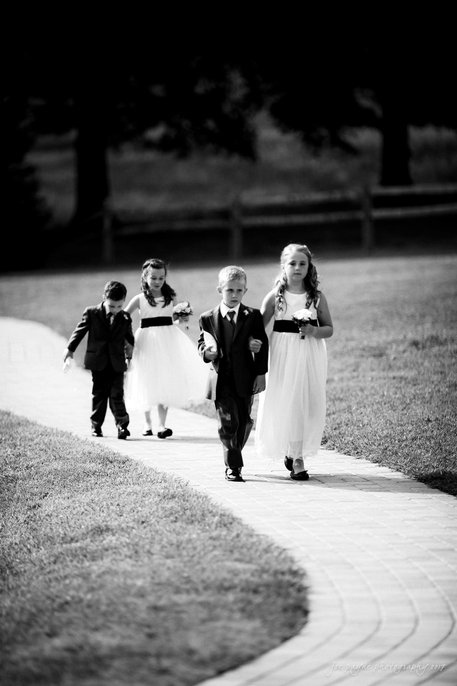 Four children in formal attire walk down a paved path, two girls in white dresses and two boys in dark suits. Theyre in a grassy outdoor setting with a fence in the background.