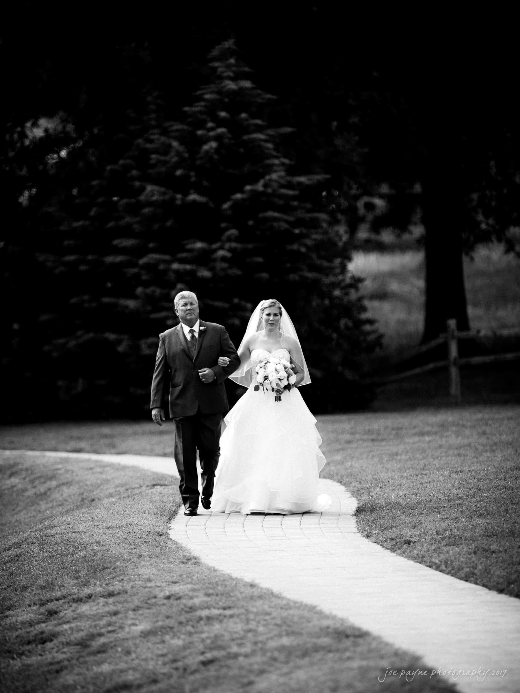 A bride in a wedding gown and veil walks down a curving stone pathway with an older man in a suit, surrounded by grass and trees. She holds a bouquet of flowers. The image is in black and white.