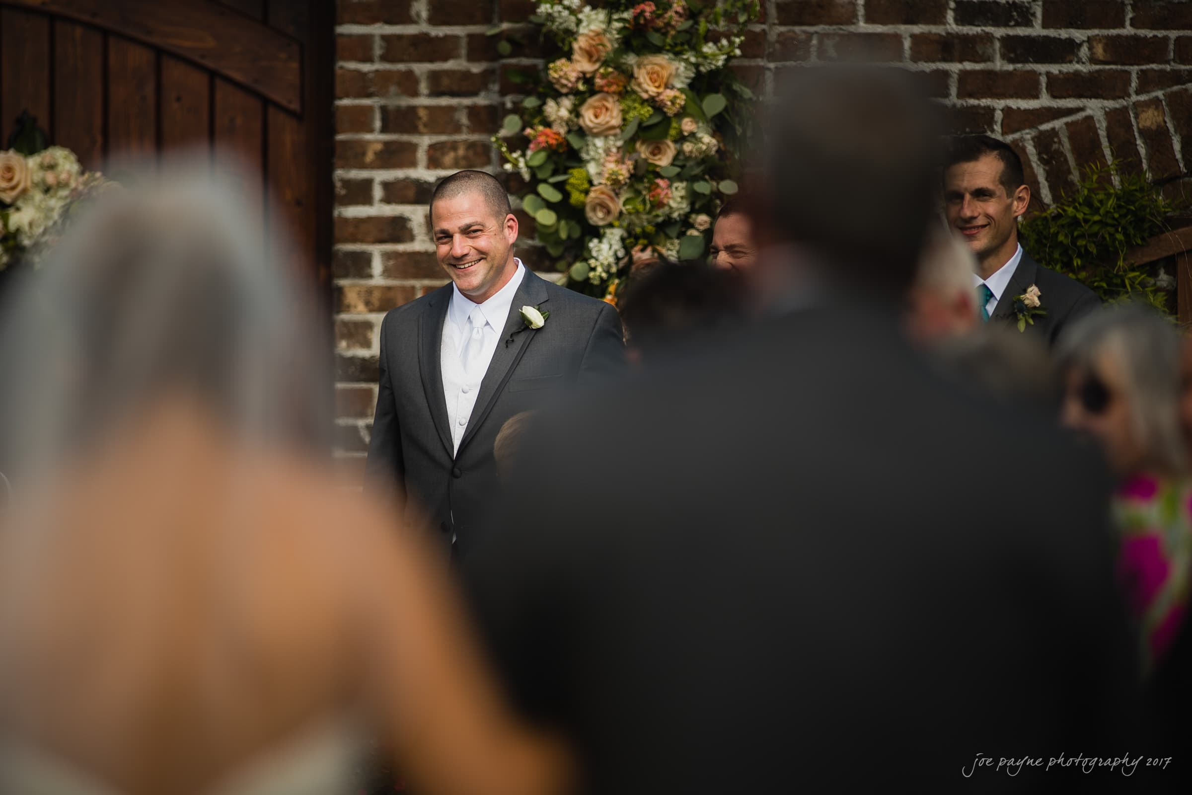 A groom in a suit smiles warmly during an outdoor wedding ceremony. Hes standing against a brick wall adorned with a floral arrangement. The blurred figures of guests are visible in the foreground.