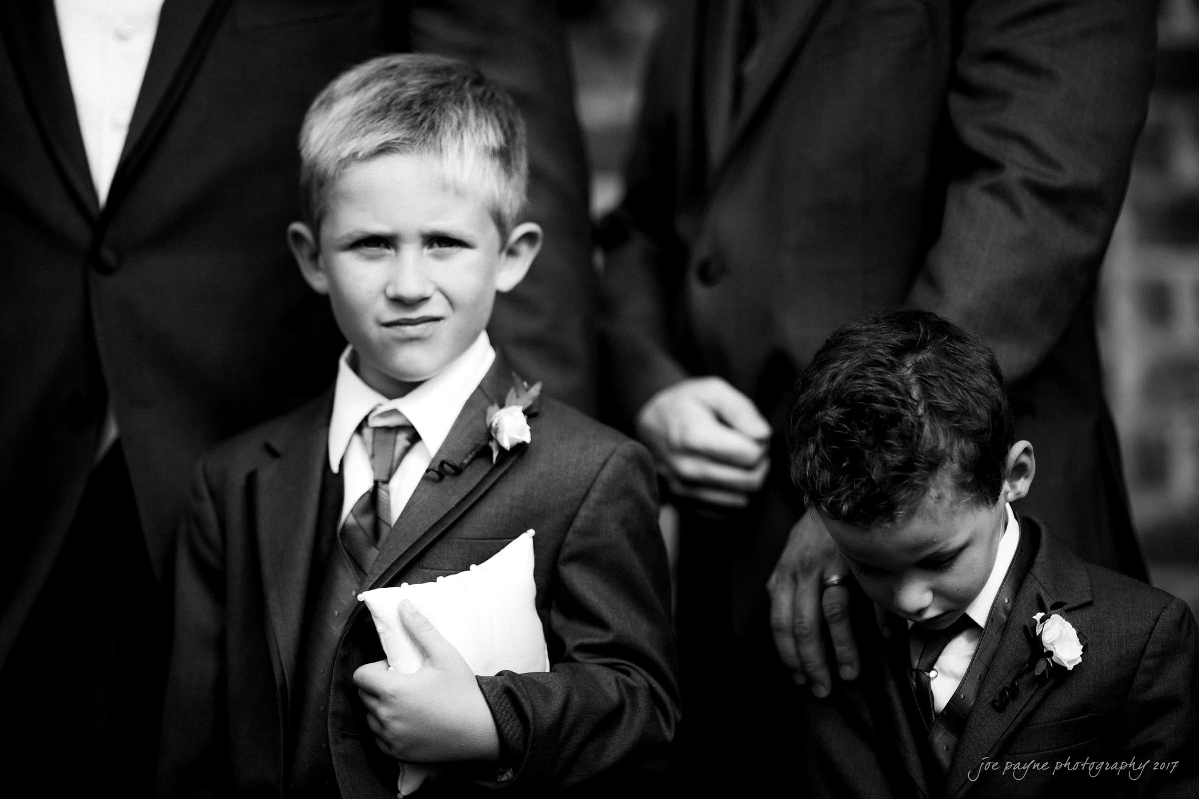 Two young boys dressed in suits stand outdoors. The boy on the left holds a cushion and looks at the camera, while the boy on the right looks down. They are surrounded by adults in suits, partially visible in the background.