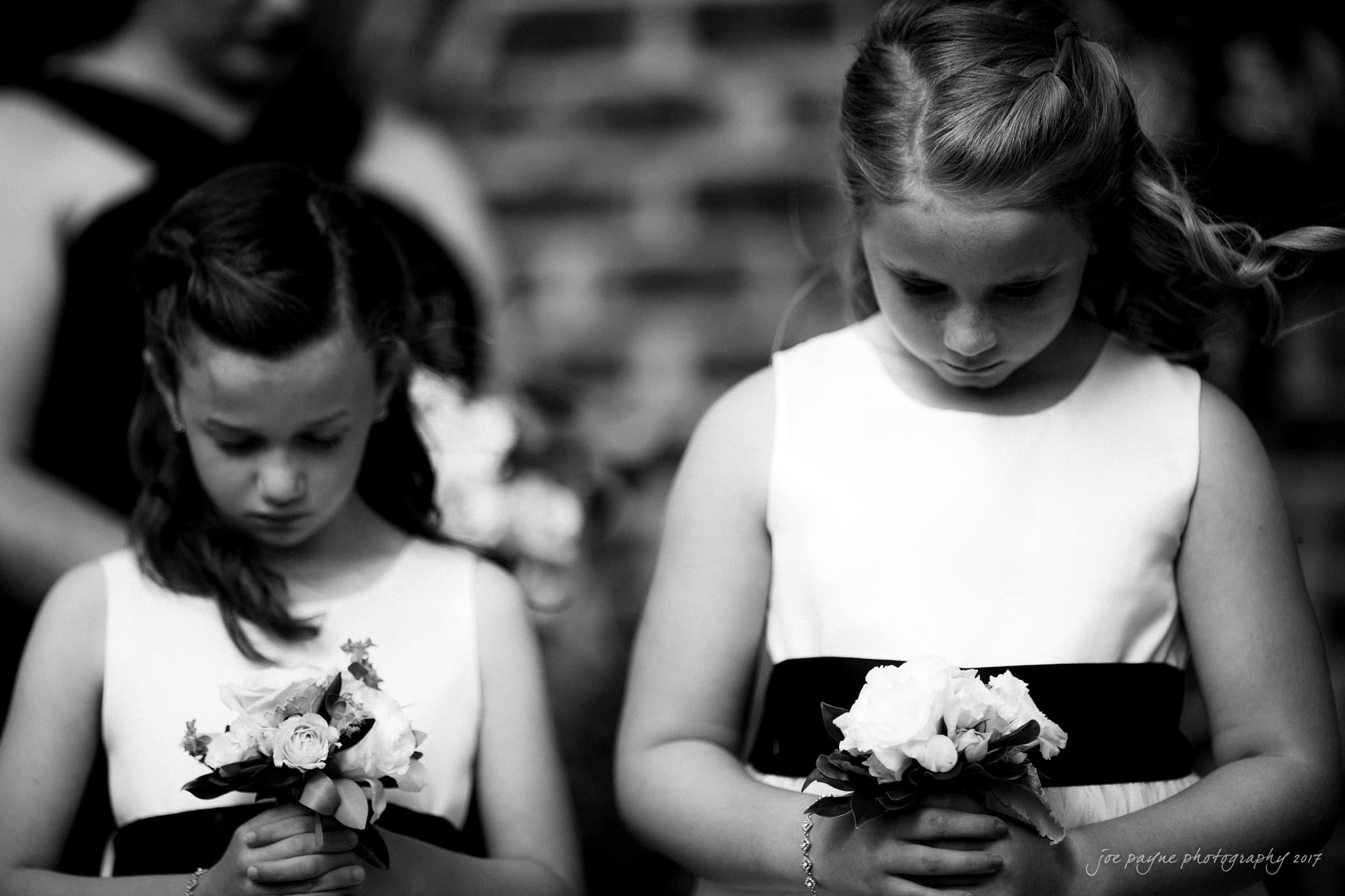 Two young girls in dresses hold bouquets, looking down solemnly. The image is in black and white, capturing a moment of quiet reflection. The background is blurred, with a hint of brick wall.