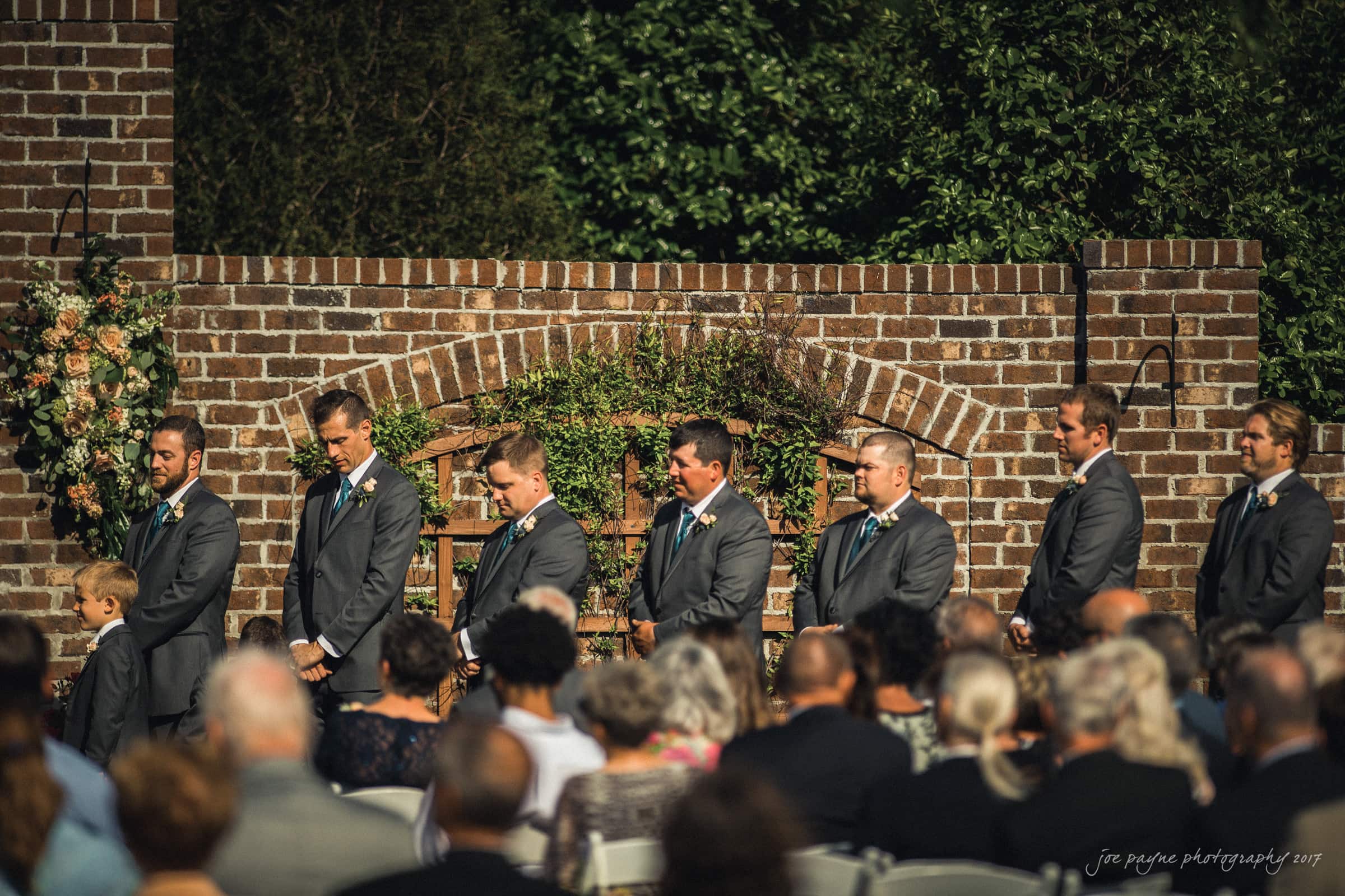 A group of groomsmen in gray suits stand in a line outdoors at a wedding ceremony. They are in front of a brick wall with floral arrangements. Guests in formal attire are seated in the foreground, with trees in the background.