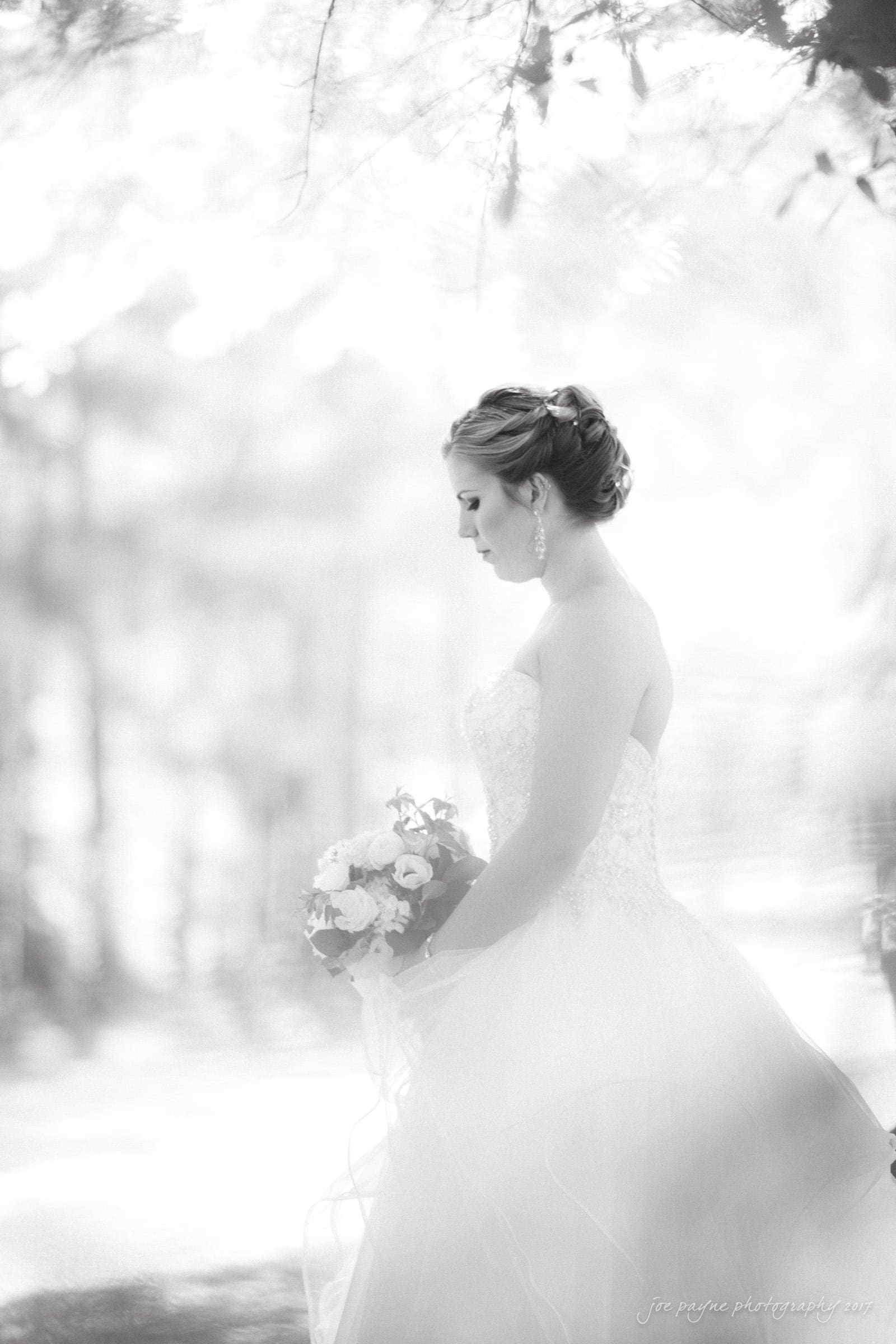 Black and white image of a bride in profile holding a bouquet. She is wearing an elegant strapless wedding gown with her hair styled up. The background is softly blurred, creating a dreamy atmosphere.
