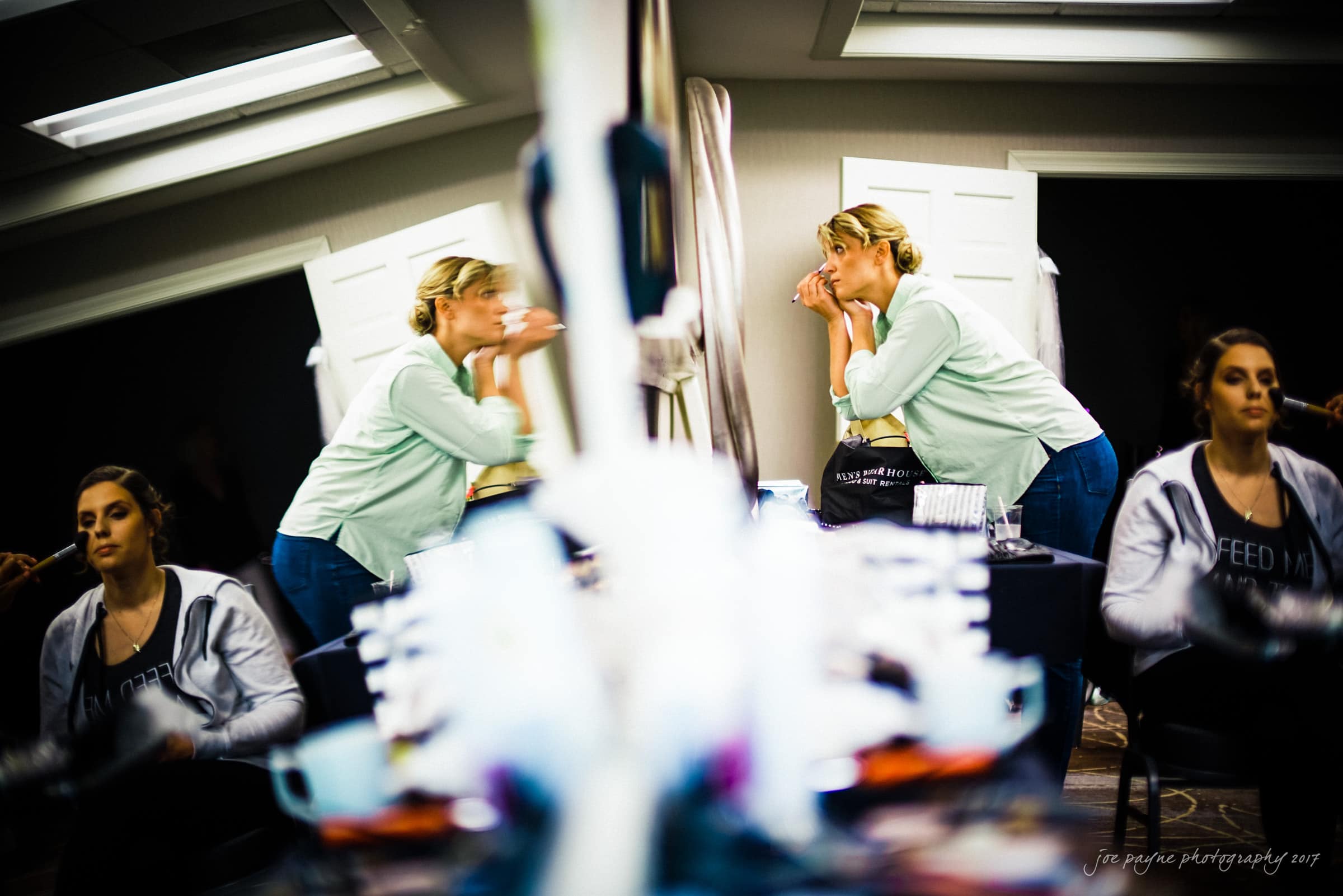 A woman applying makeup at a vanity, reflected in a mirror. Shes in a light-filled room, and the vanity is cluttered with various cosmetic items. Another woman, partially visible, sits nearby.