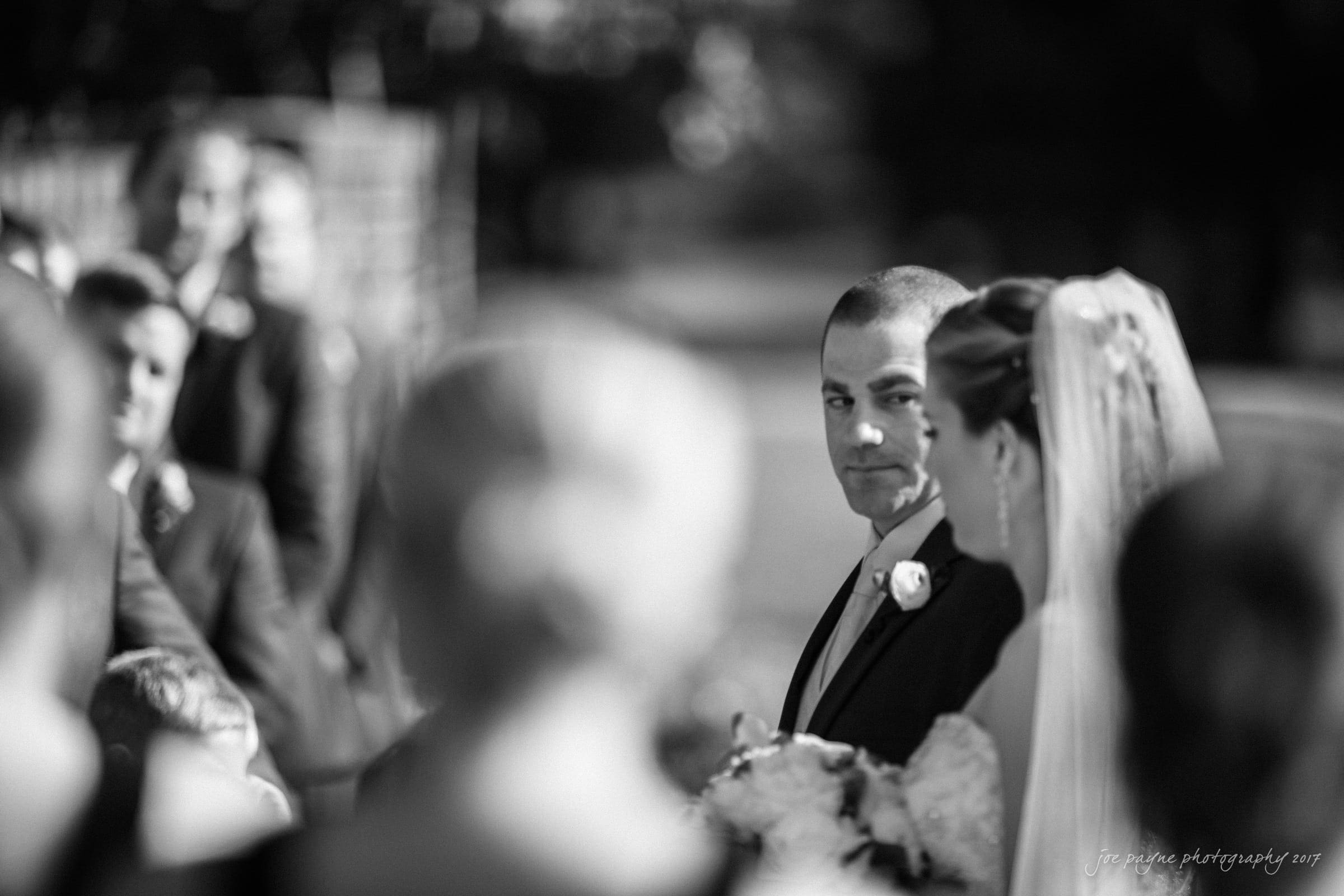 A groom in a suit stands beside a bride in a wedding dress during an outdoor ceremony. The scene is in black and white, with guests blurred in the foreground, capturing an emotional moment between the couple.
