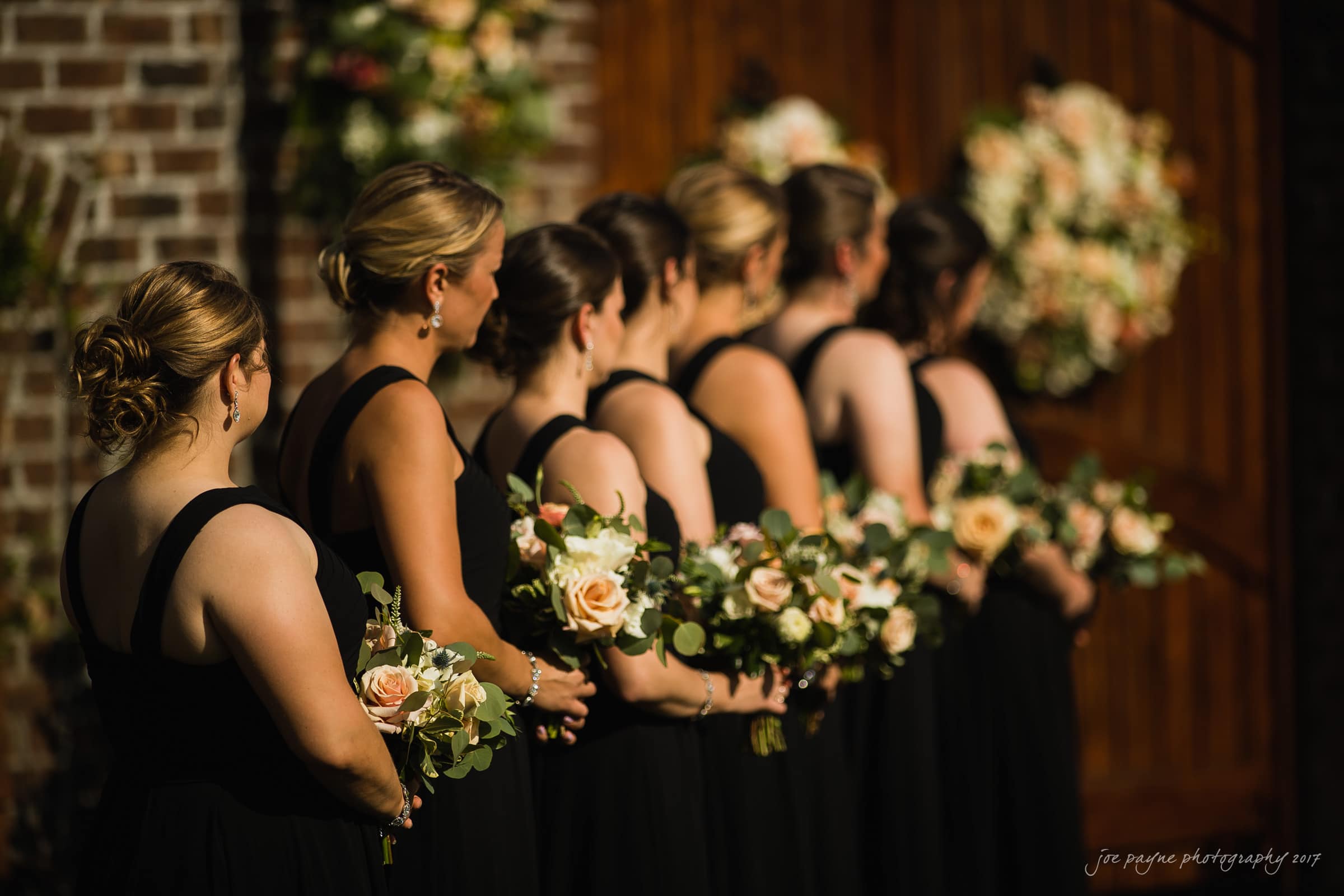 A group of bridesmaids in matching black dresses hold bouquets of flowers with white and pink roses. They stand in a row outdoors, with warm sunlight illuminating the scene. A floral arrangement decorates the wooden background.