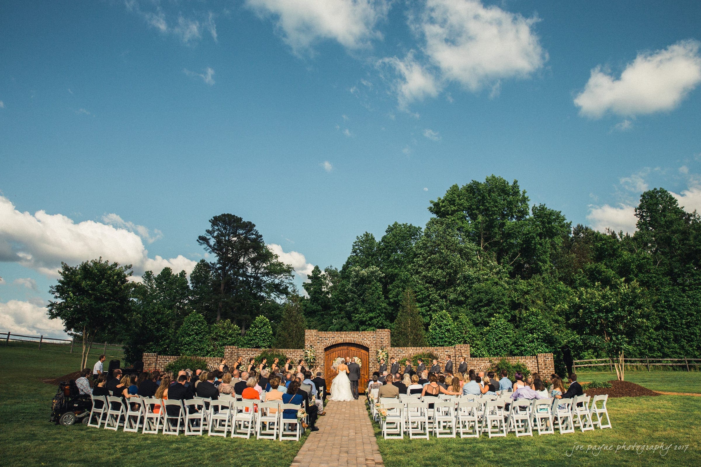 Outdoor wedding ceremony with a couple standing at an altar framed by a stone archway and surrounded by lush greenery. Guests are seated on white chairs under a bright blue sky with fluffy clouds.