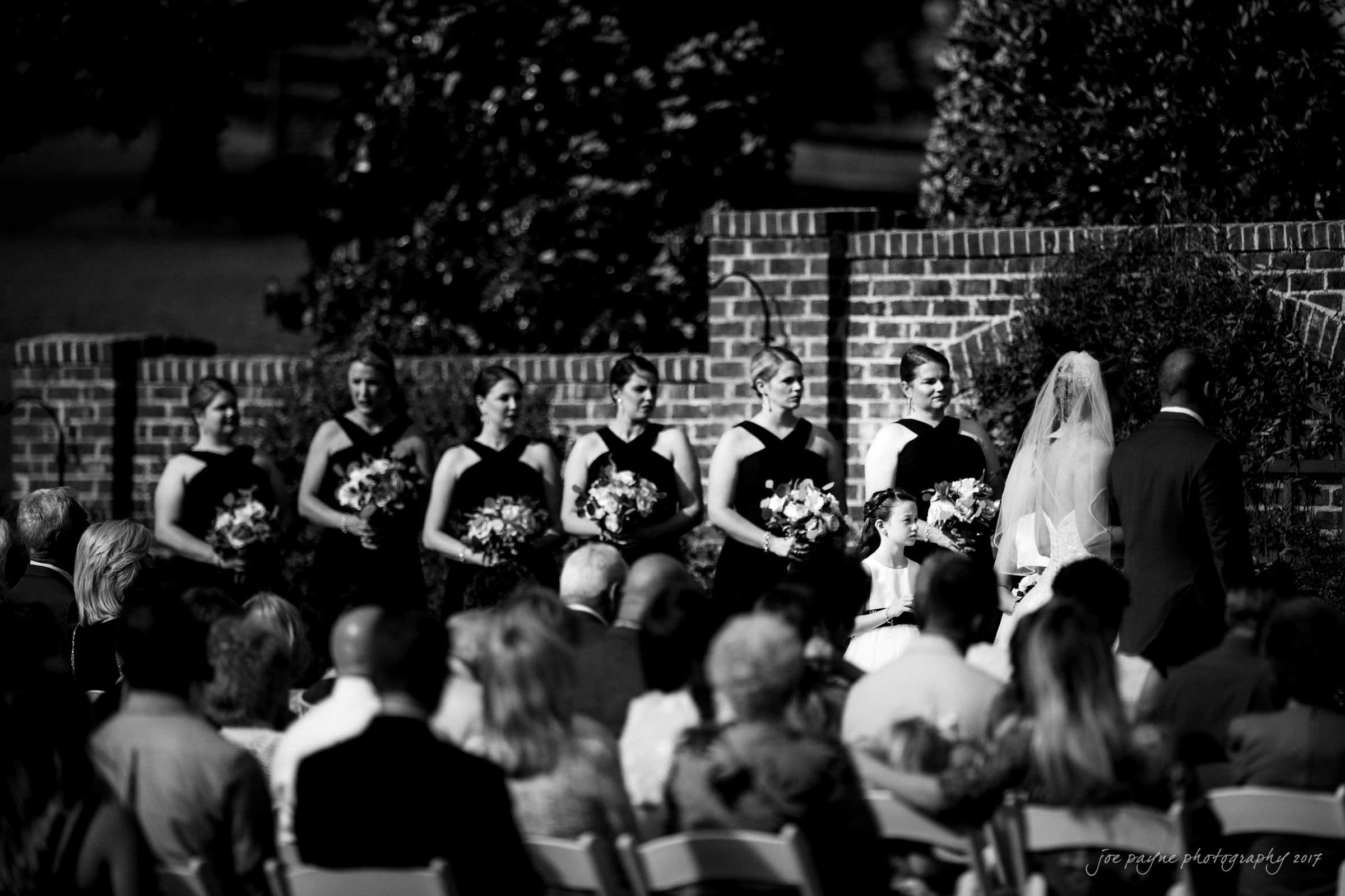 A black and white photo of a wedding ceremony outdoors. Bridesmaids in black dresses stand in a row holding bouquets, while a bride and groom, accompanied by a flower girl, face each other. Guests are seated in the foreground.