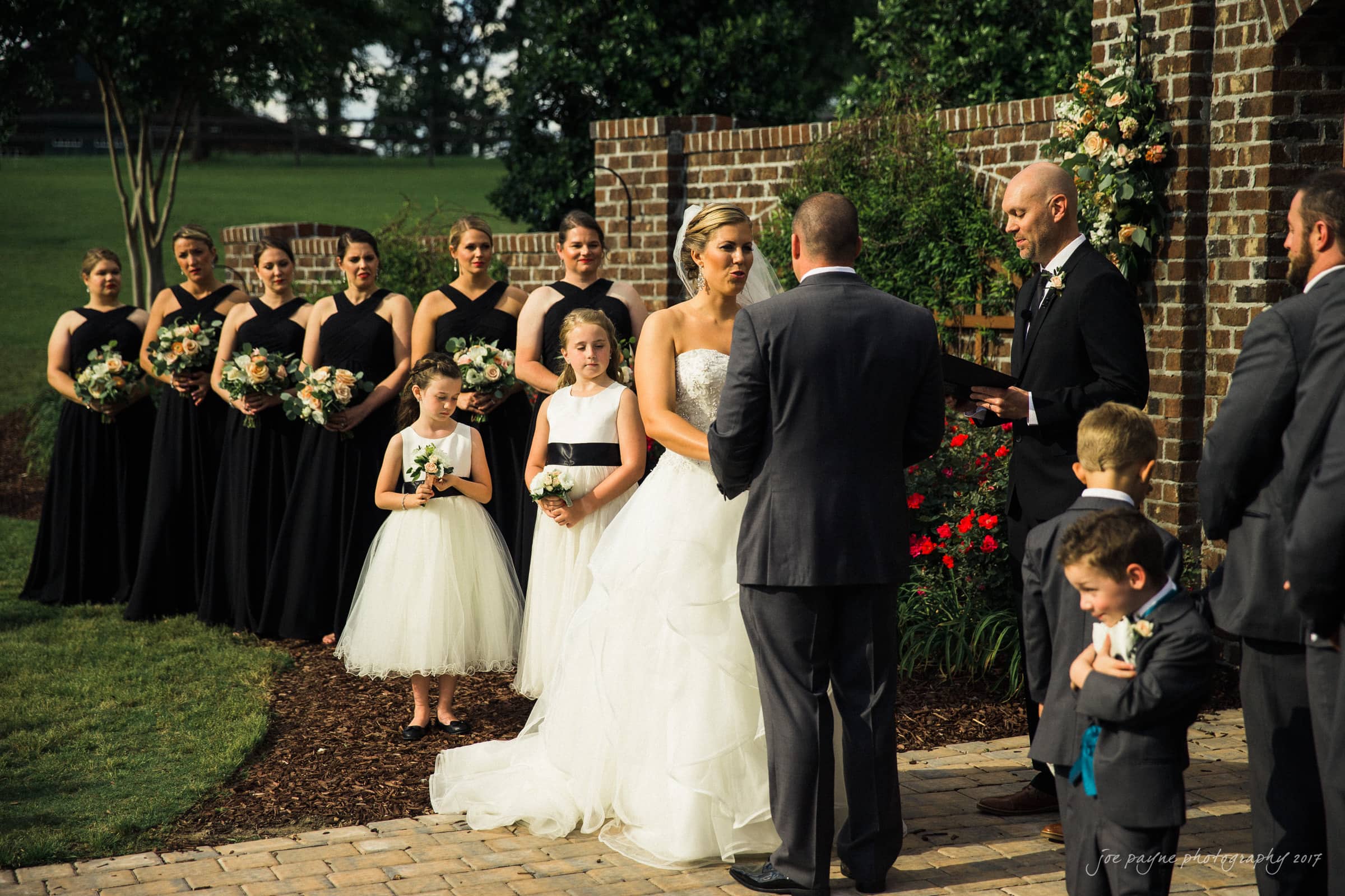 A wedding ceremony outdoors with a bride in a white gown and a groom in a suit standing before an officiant. Bridesmaids in black dresses stand to the side, and children are present. The scene is set against a brick wall with greenery.