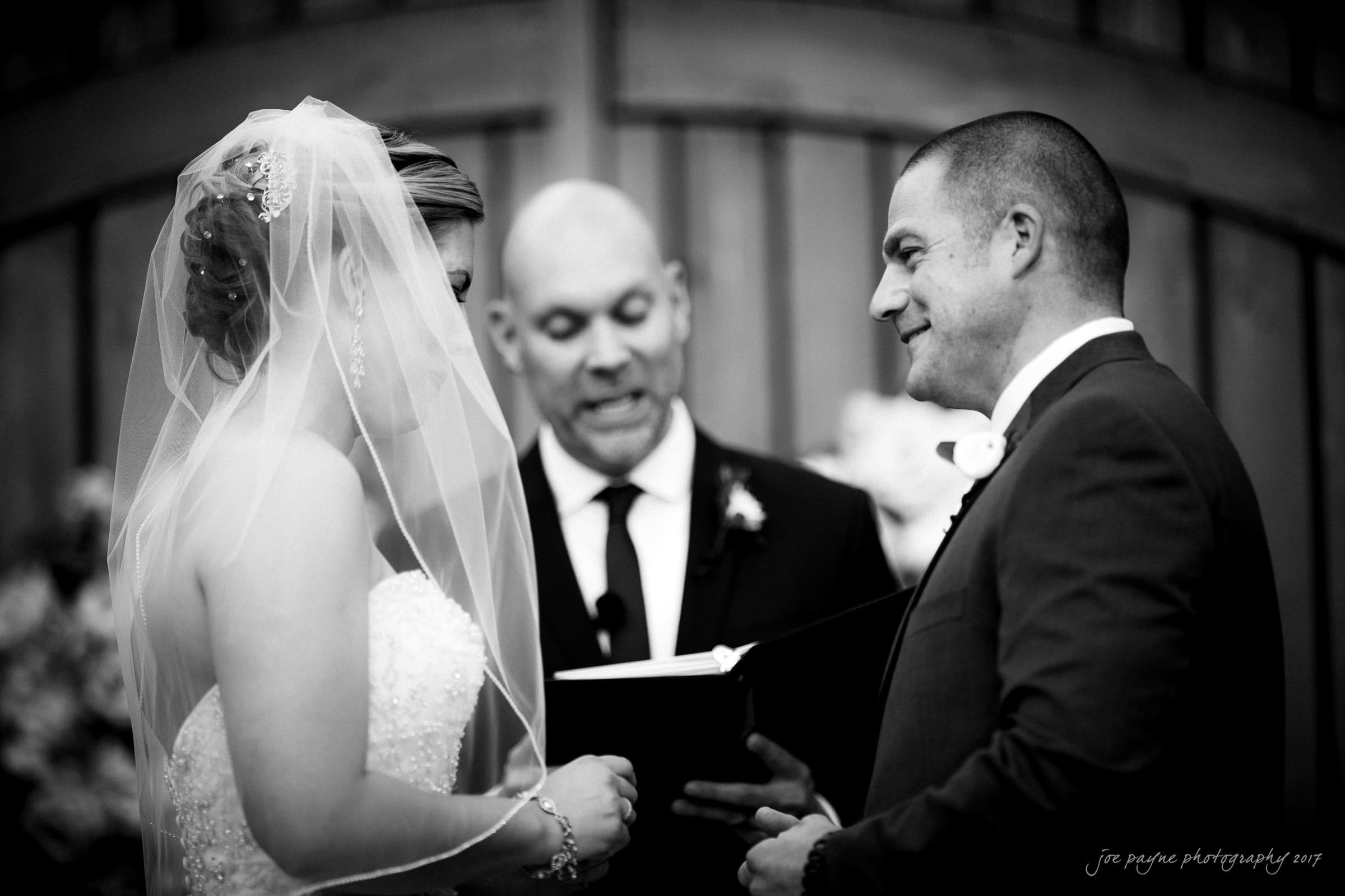 A bride and groom stand facing each other during a wedding ceremony. The bride wears a veil and a strapless dress, while the groom is in a suit. A celebrant stands between them, holding a book. The background features wooden doors. The photo is in black and white.