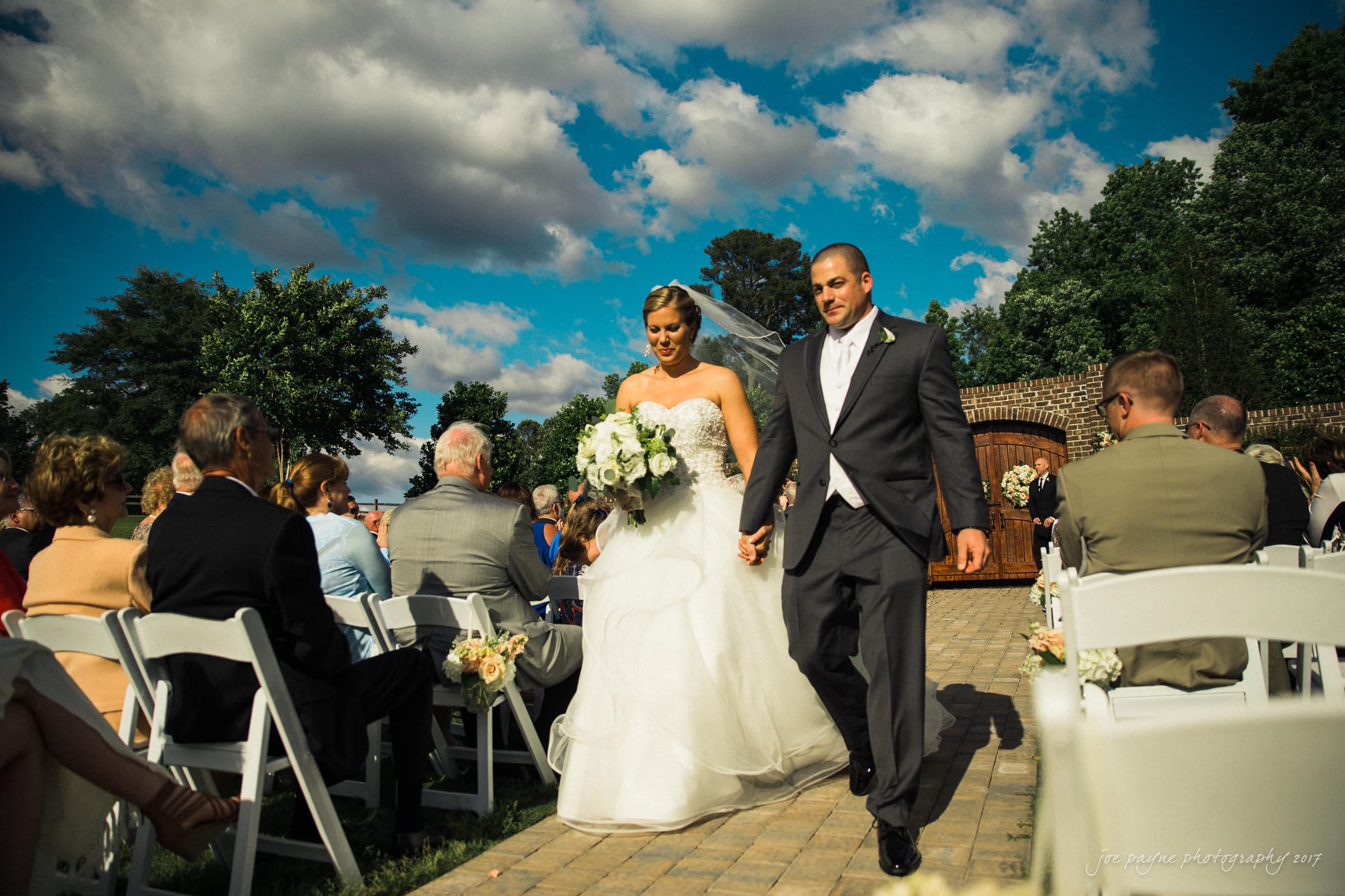 A bride and groom walk down an outdoor aisle hand in hand, surrounded by seated guests. The bride holds a bouquet of white flowers, and both wear formal attire. The sky is partly cloudy with a few trees in the background.