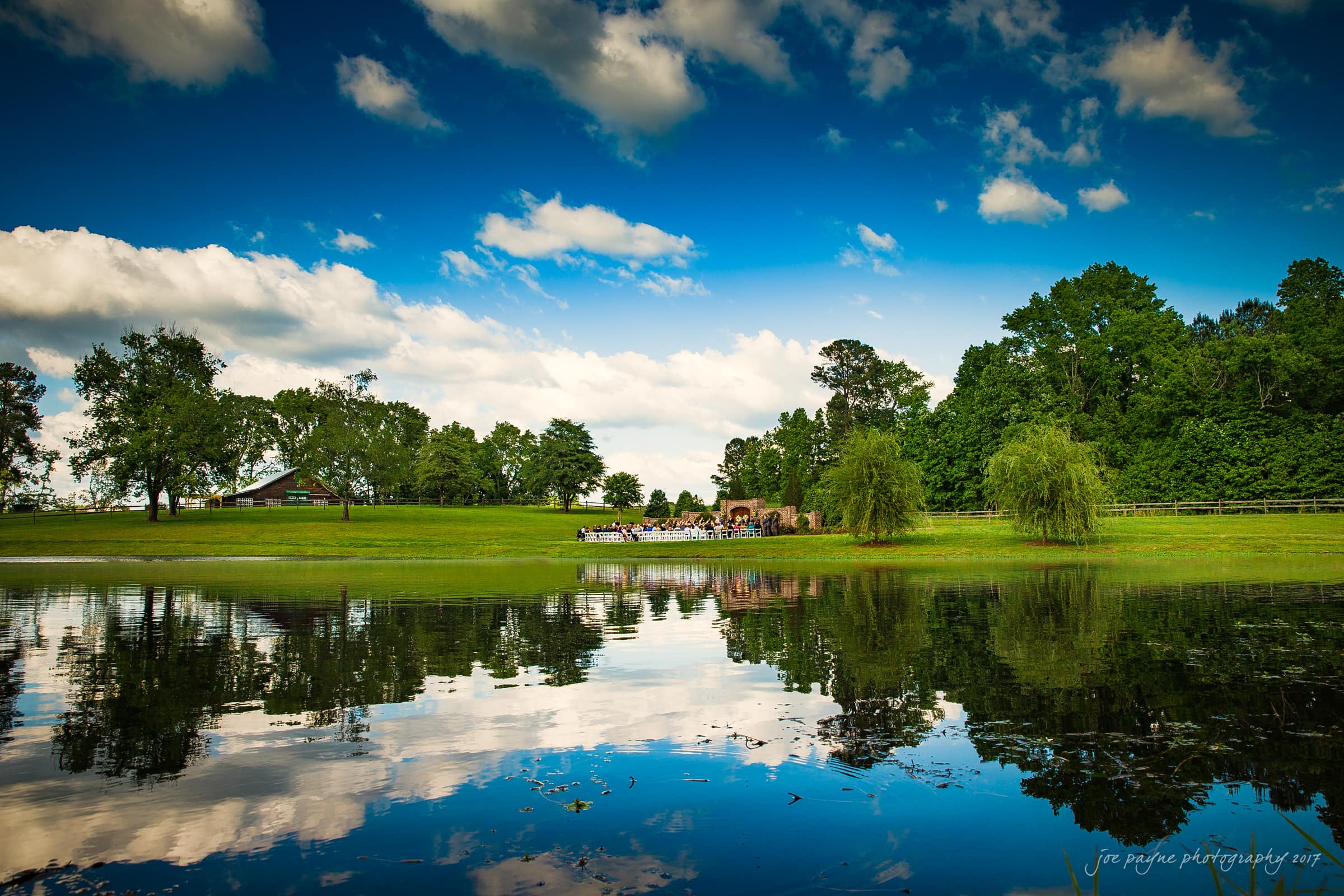 A serene landscape with a pond reflecting a bright blue sky and fluffy clouds. Grassy fields and lush green trees surround the pond, and a small gathering is visible in the distance, near a wooden building.