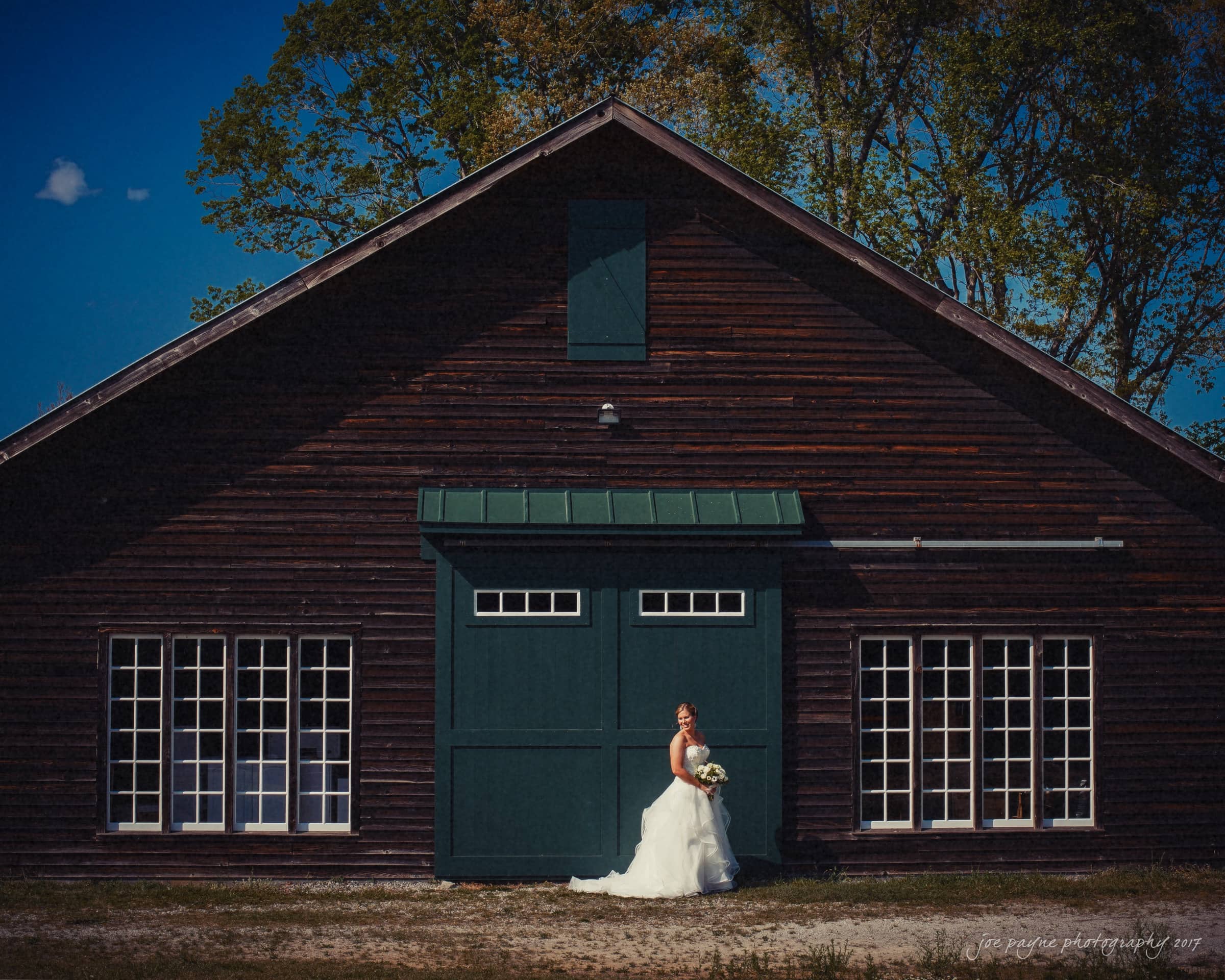 A bride in a white gown stands holding a bouquet in front of a rustic wooden barn with large green doors and multiple windows. The barn is set against a backdrop of tall trees under a clear blue sky.