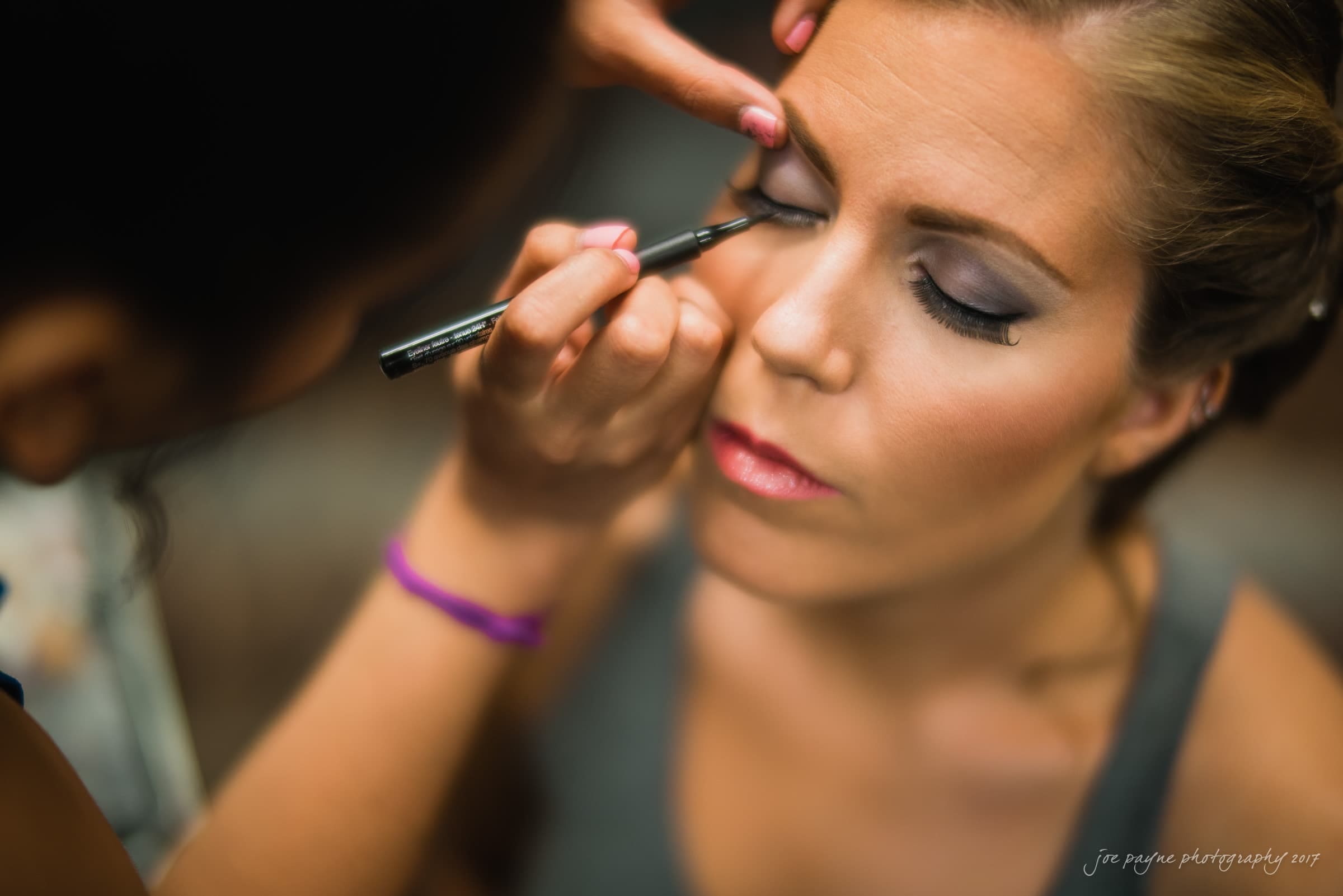 A woman with closed eyes is having eyeliner applied by another person. She is wearing subtle pink lipstick and her hair is tied back. The focus is on her face, highlighting the makeup application process.