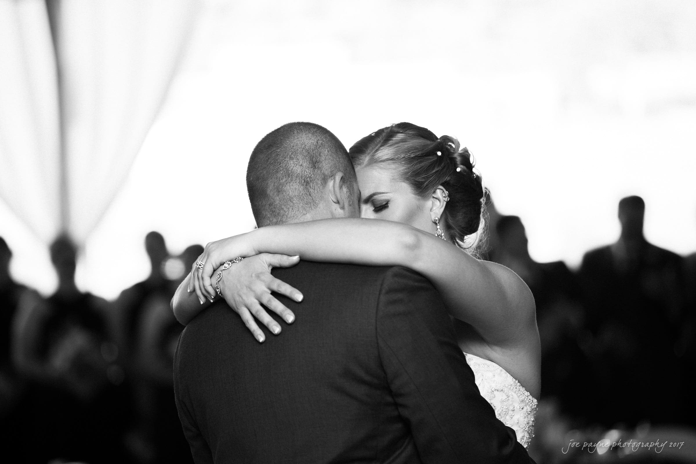 A couple is dancing closely at their wedding. The bride, in a strapless gown, has her arms around the groom, who is wearing a suit. The black and white photo captures a tender moment with blurred guests in the background.