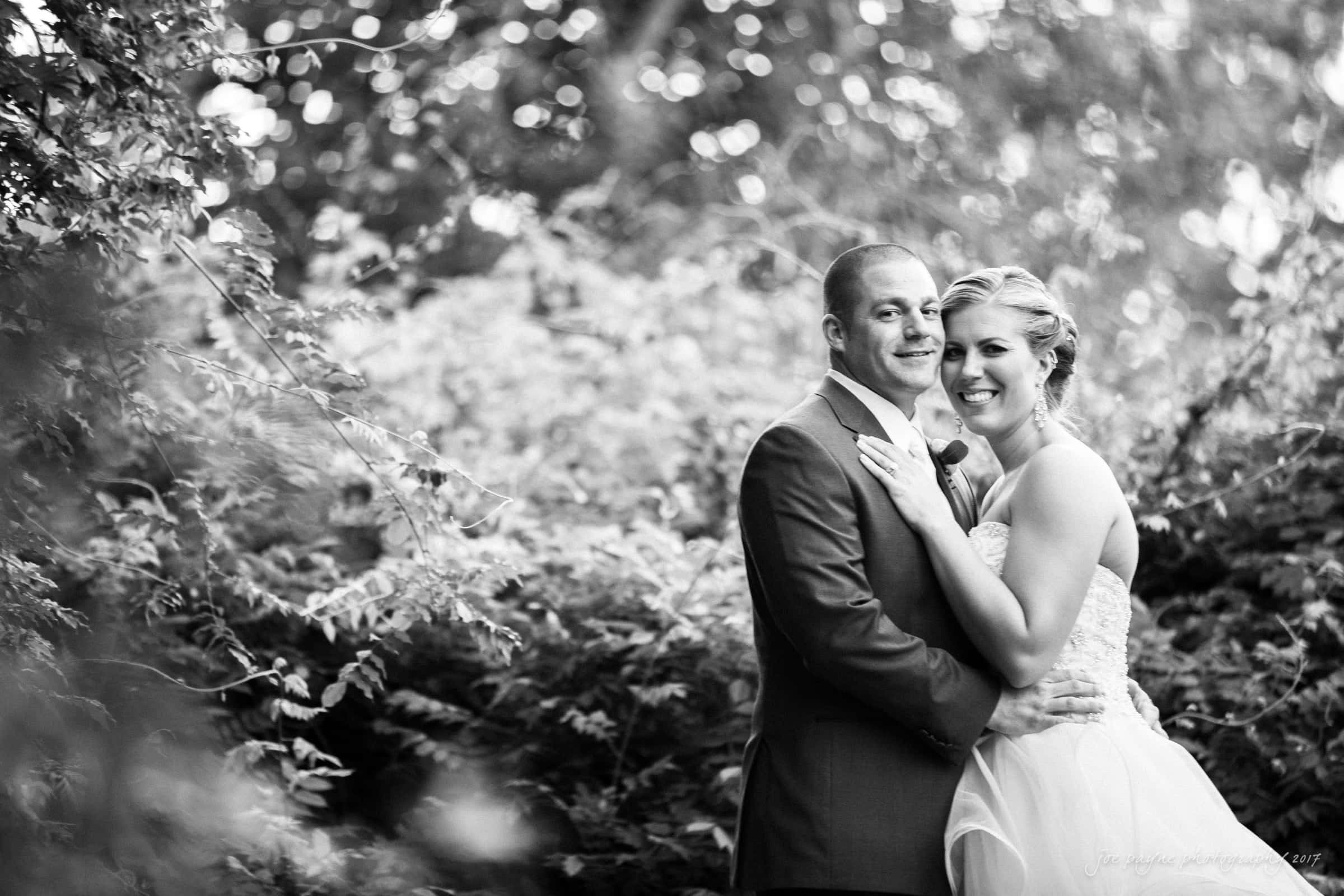 A black and white photo of a couple in wedding attire, standing close together and smiling amidst lush foliage. The groom is in a suit, and the bride is in a strapless gown. Sunlight filters through the trees in the background.