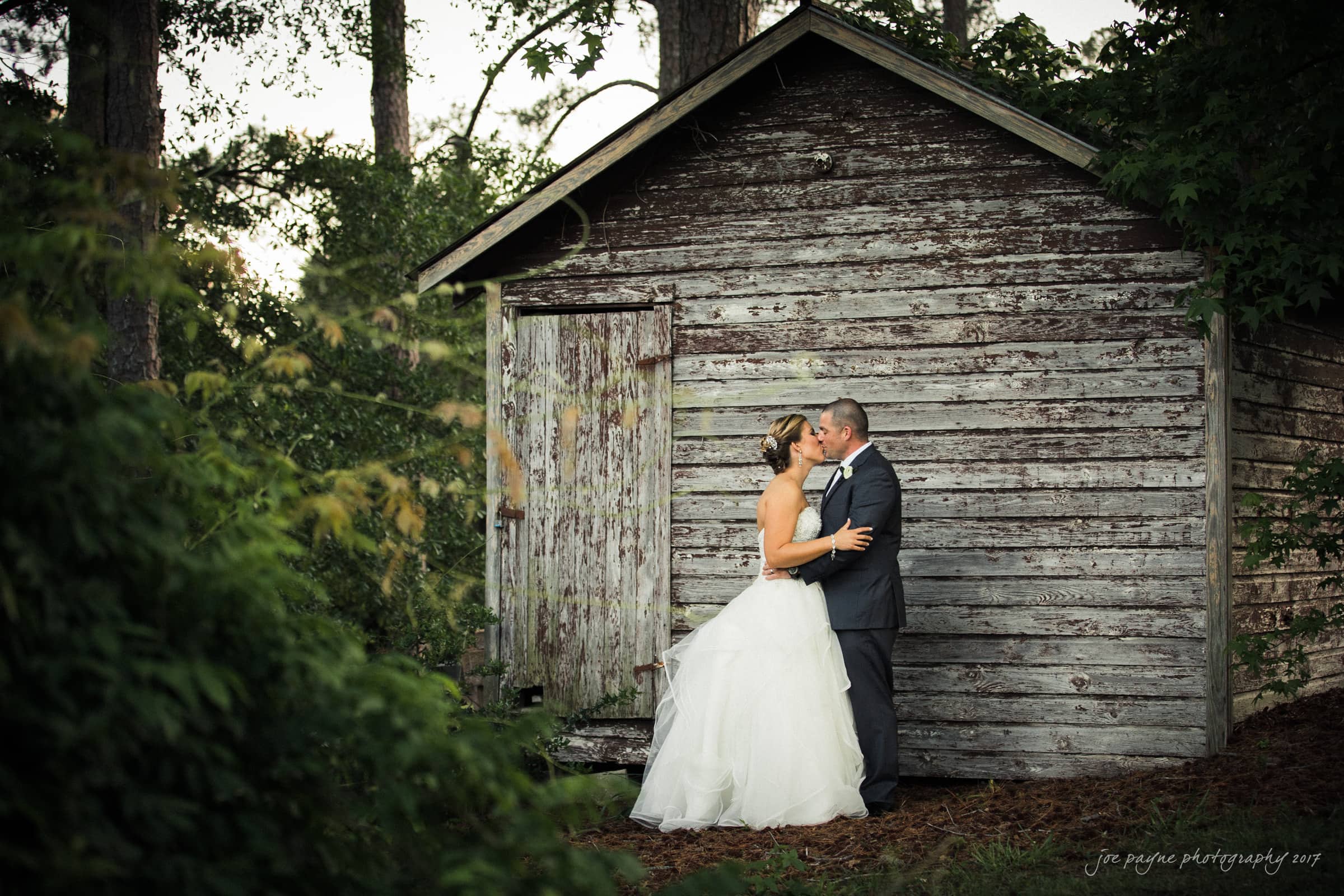 A bride and groom embrace lovingly in front of a rustic wooden shed, surrounded by lush green foliage and trees, creating a serene and romantic outdoor setting.