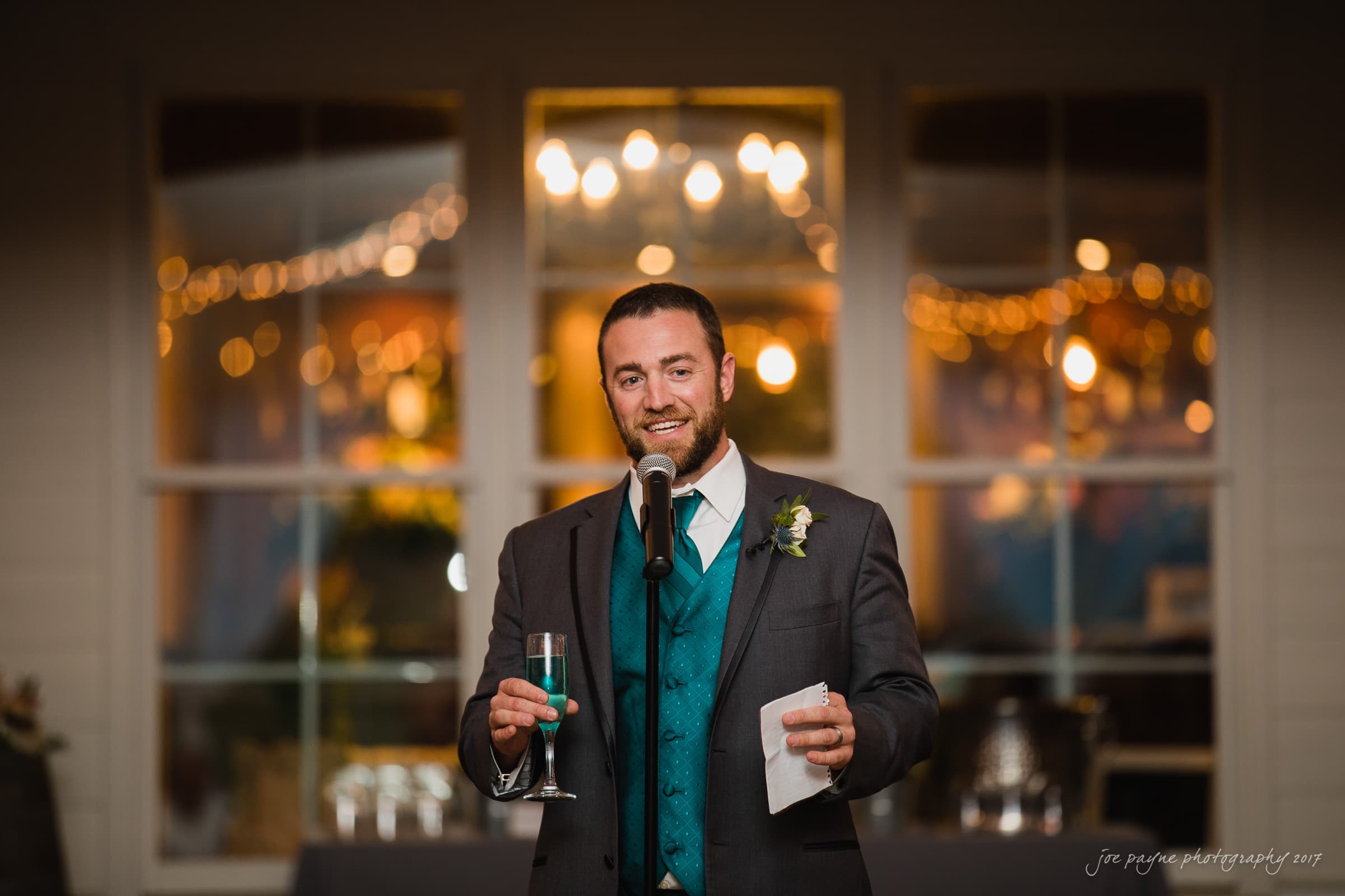 A man wearing a suit and teal vest stands by a microphone, holding a champagne glass and paper, appearing to make a speech. Warm, decorative lights and blurred background create a festive atmosphere.