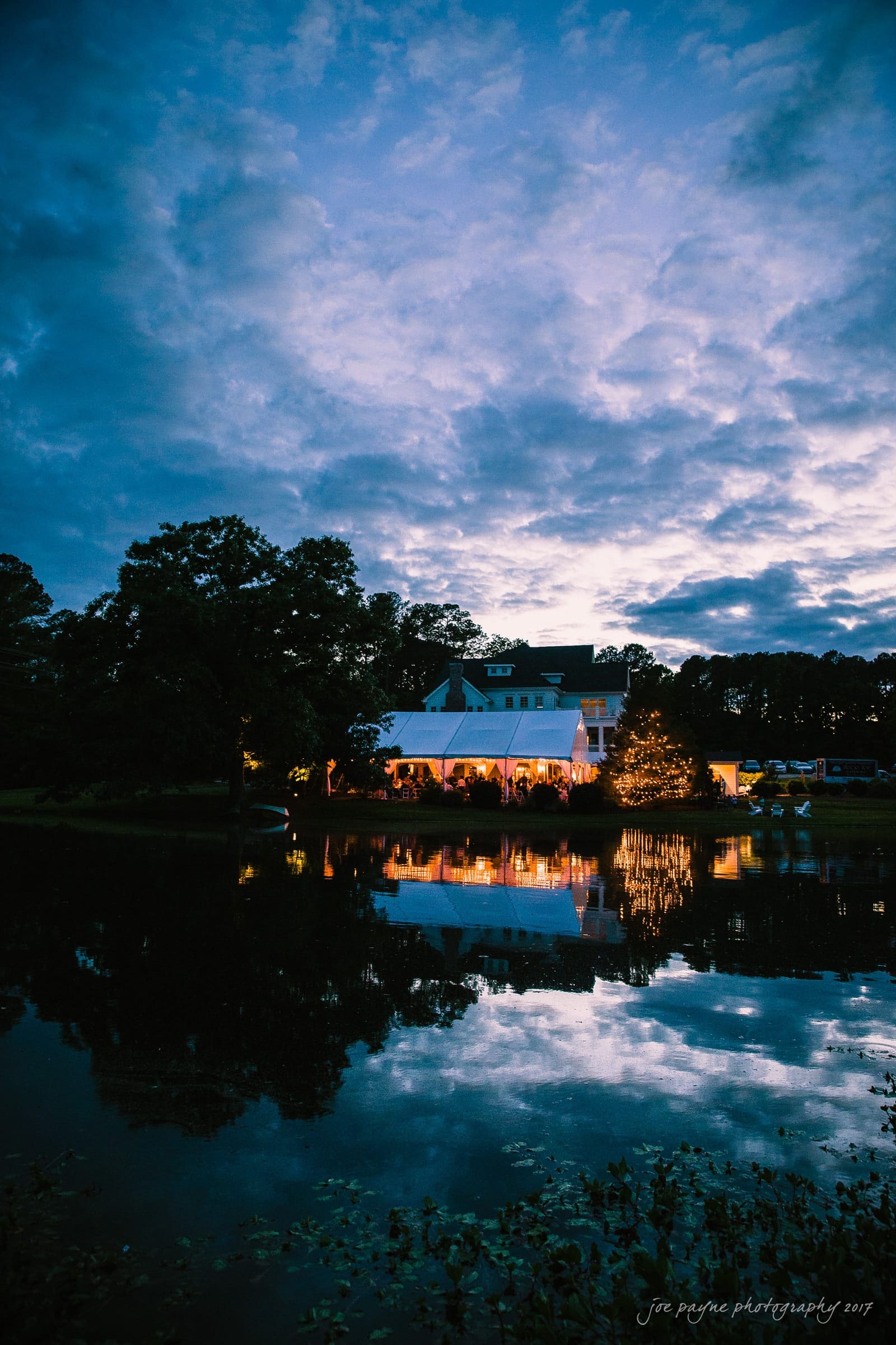 A large house with glowing windows is reflected in a calm lake at dusk. The sky is filled with dramatic clouds, and trees frame the scene, creating a serene and picturesque evening landscape.