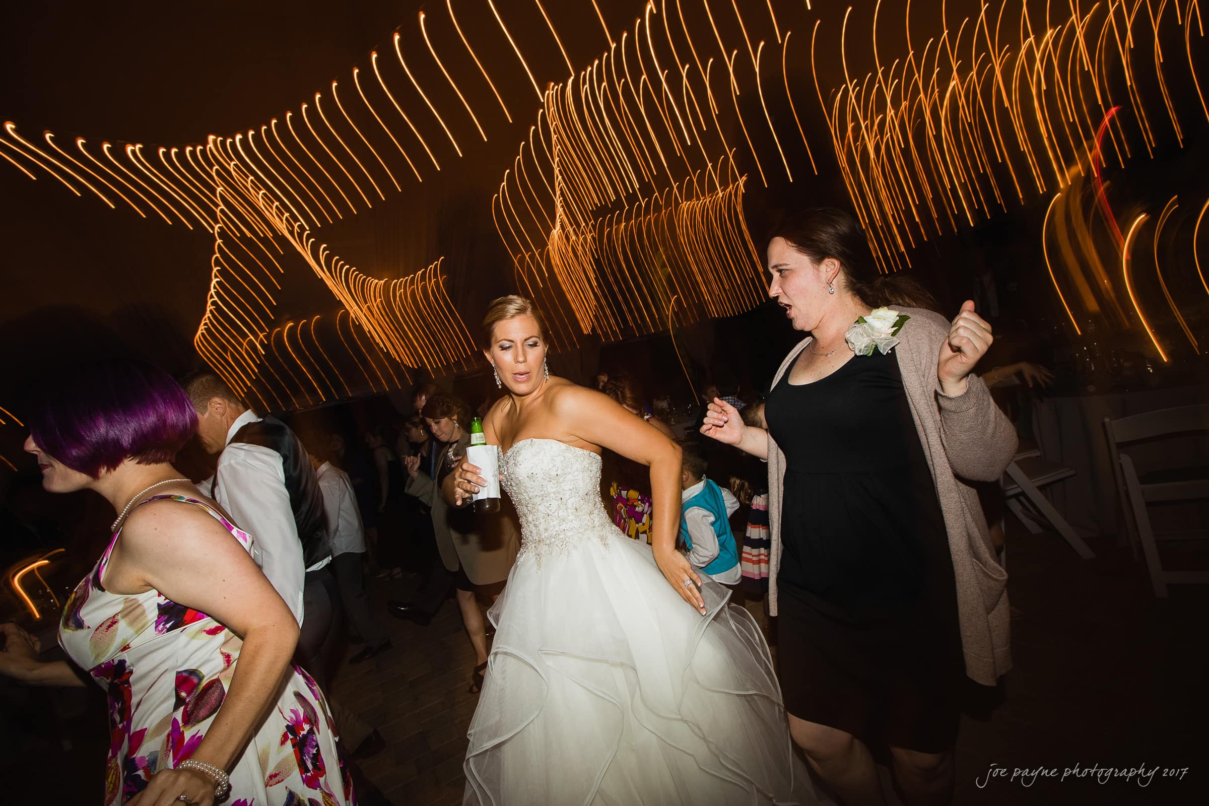 A bride in a white dress dances energetically with guests at an indoor reception. Motion blur from lights creates a dynamic atmosphere. Several people are visible, engaged in the lively celebration.