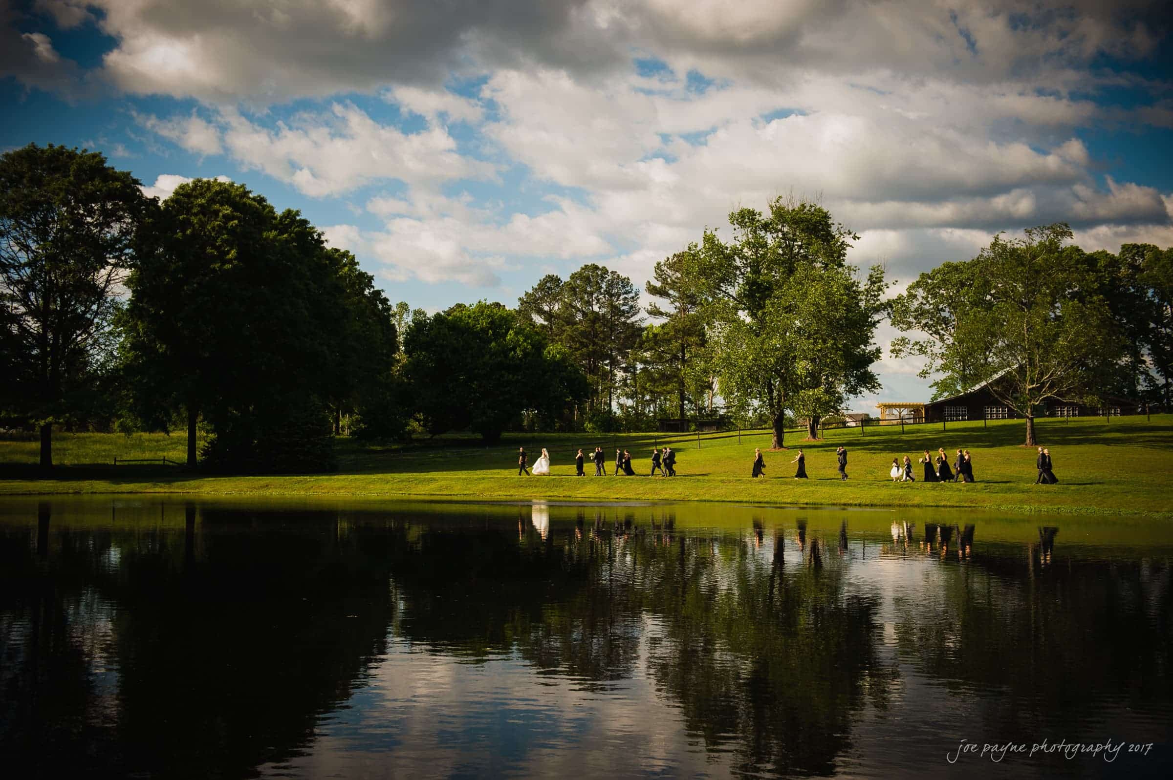 A group of people in formal attire walk in a line next to a serene lake, surrounded by lush green grass and trees. The sky is partly cloudy, and there is a pavilion in the background. The scene is reflected in the lakes calm water.