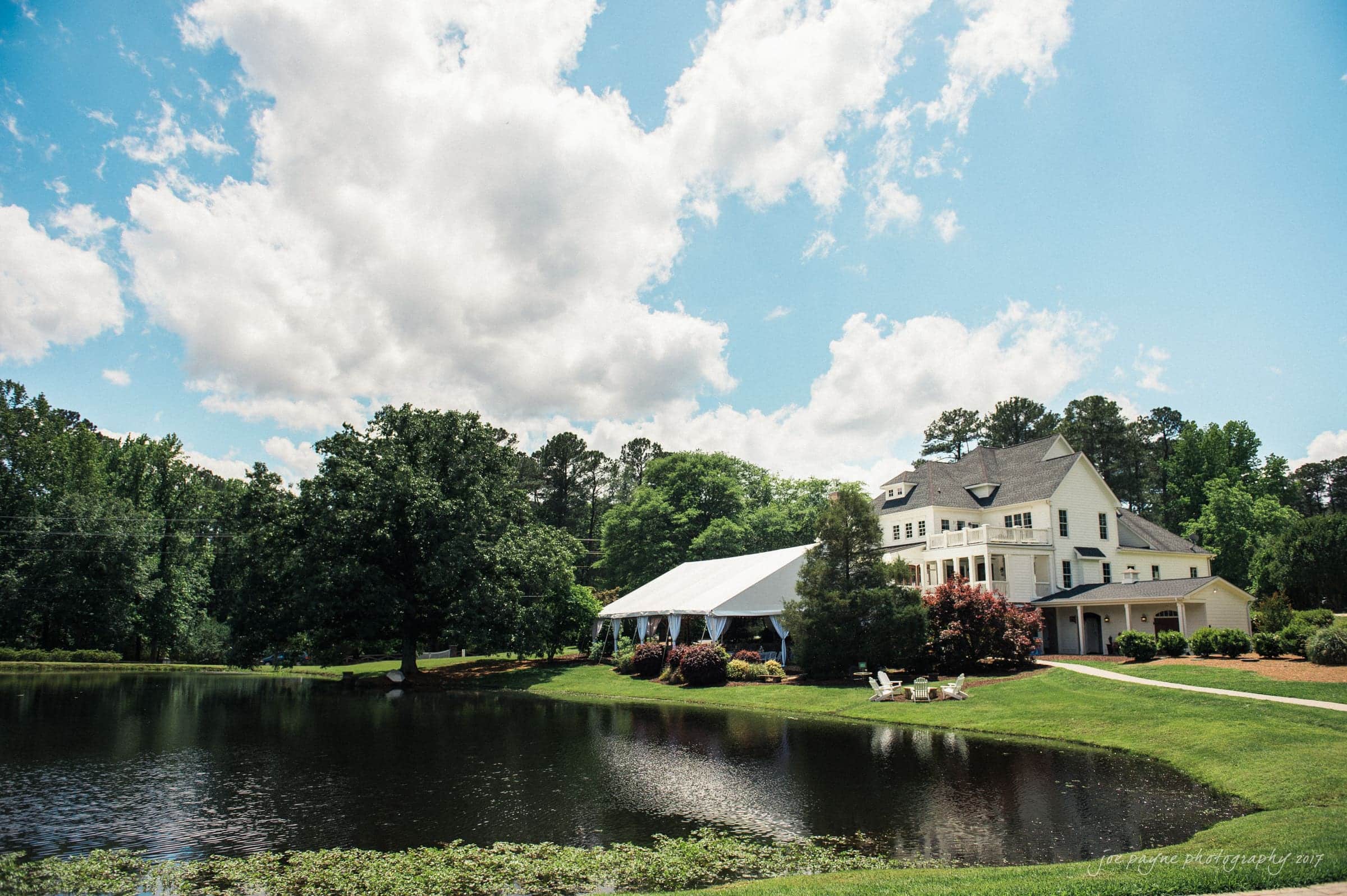 A picturesque scene of a large house with a tent beside it, set against a backdrop of lush green trees and a cloudy blue sky. A small pond reflects the surrounding landscape, adding tranquility.