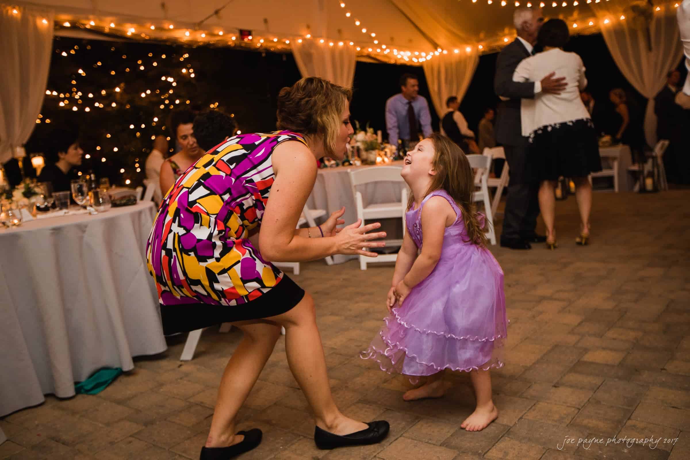 A woman and a young girl in a purple dress joyfully dance together at an outdoor event. The area is decorated with string lights and draped curtains, with people mingling and tables set with dishes in the background.