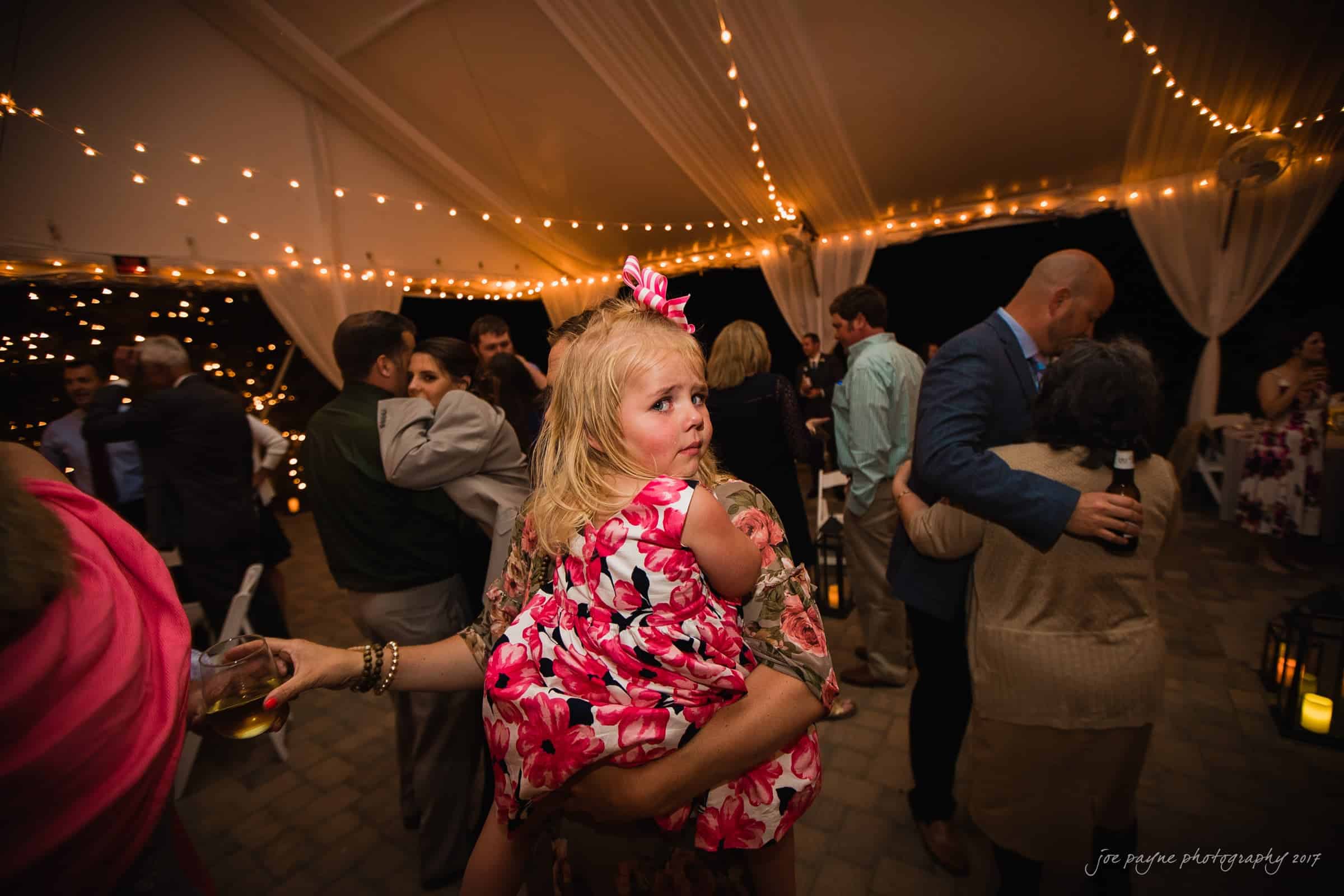 A little girl in a floral dress is carried by a woman at an outdoor event under string lights. Several people are dancing in the background, and the atmosphere appears festive and lively.